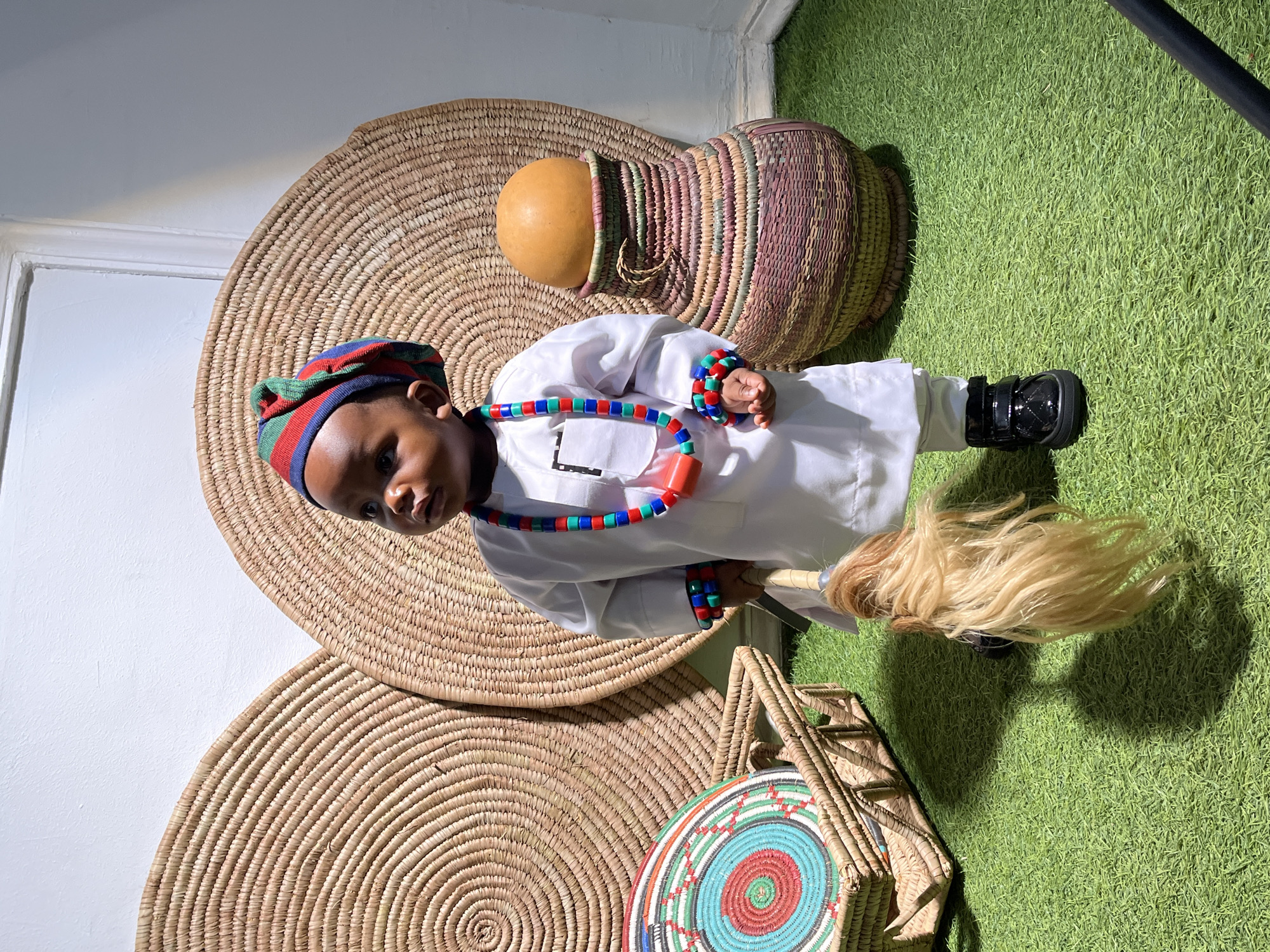 A young child, possibly a toddler, stands on artificial grass in an indoor setting. The child is dressed in a white traditional outfit, complete with a multi-colored cap and beaded necklaces and bracelets. They are holding a whisk-like object with a blond mane.
Behind the child are two large, circular woven mats displayed against a white wall. To the child's left is a woven basket containing a circular woven mat with concentric rings of red, turquoise, and white. To the child's right is a tall, woven vase with a gourd on top. The lighting suggests an indoor studio or event space. The location of Jos, Nigeria, is not visually discernible from the image itself.