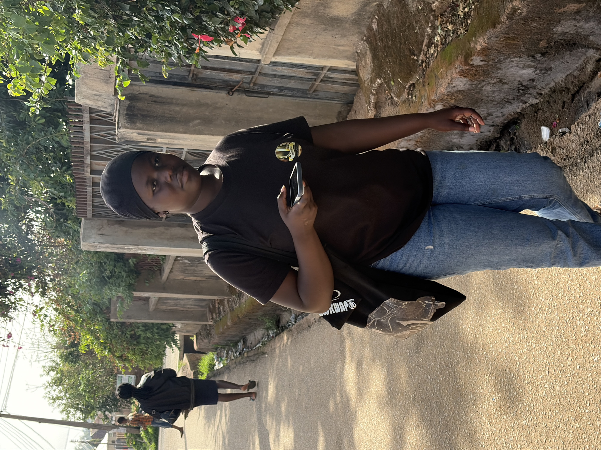 A person wearing a black head covering, a dark t-shirt, and blue jeans stands on a paved street in Jos, Nigeria. They hold a smartphone in their right hand. A black tote bag hangs from their left shoulder. Another person in a dark outfit walks in the background. The street is lined with buildings and vegetation. Debris is visible along the side of the street.
