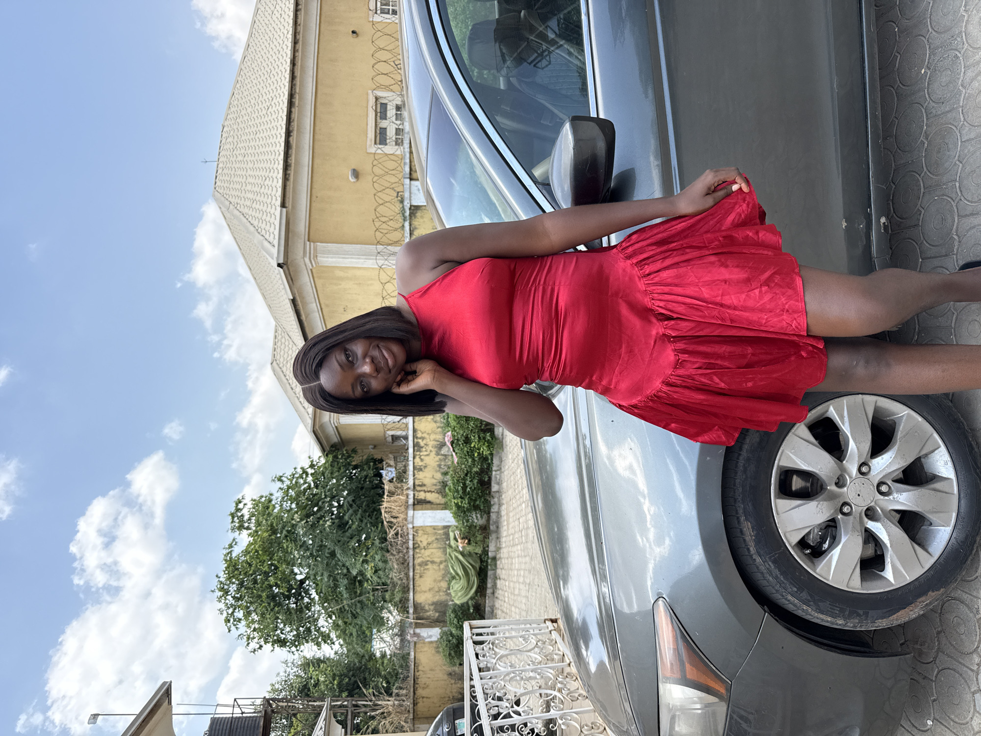 A dark-skinned woman in a short, red dress poses leaning against a grey car. She smiles, her right hand to her face. The car is parked on a paved surface near a building with a light-yellow wall and white tiled roof. Green trees and shrubs grow along a fence in the background. The sky is blue with scattered white clouds. The scene is set outdoors in what appears to be a residential area in Abuja, Nigeria, indicated by the style of buildings and environment.