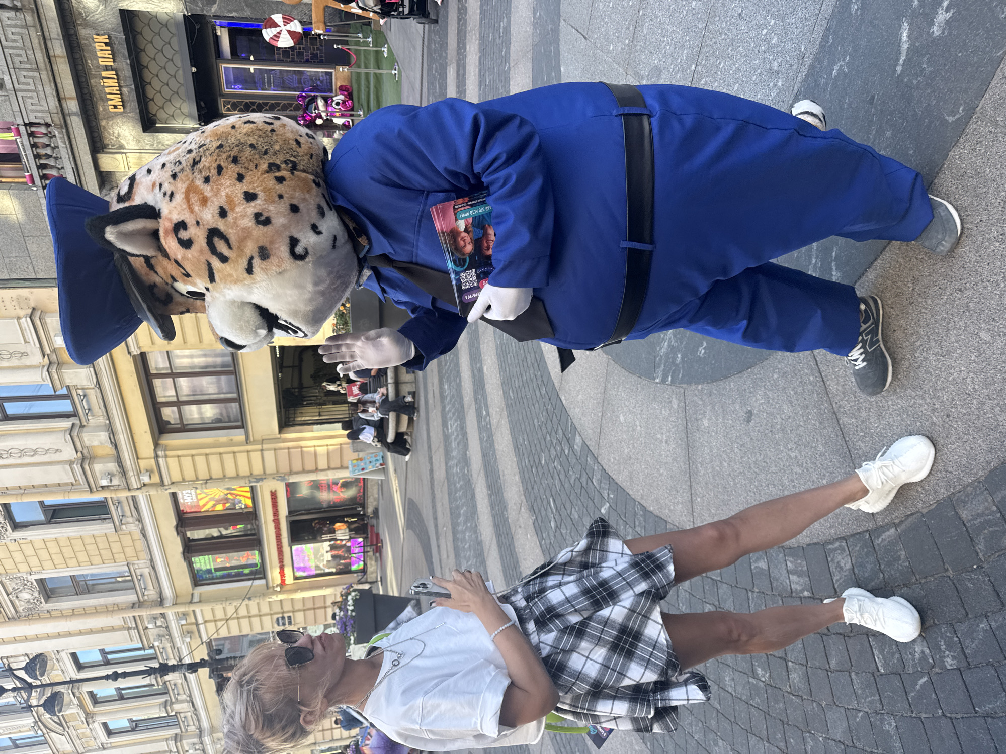In Saint Petersburg, Russia, a person in a leopard mascot costume, wearing a blue jumpsuit and white gloves, interacts with a woman. The mascot holds a colorful magazine in its hand. The woman, dressed in a white t-shirt and a black and white plaid skirt, wears sunglasses and jewelry. She holds a smartphone in her hand. The scene is outdoors on a paved area with a patterned walkway. In the background, buildings with ornate architecture line the street. Some buildings have illuminated signs and storefronts. The time of day appears to be daytime, with sunlight casting shadows. The weather seems pleasant. A sign visible on a building reads "СМАЙЛ ПАРК" (Smile Park). The overall atmosphere suggests a public gathering or promotional event.