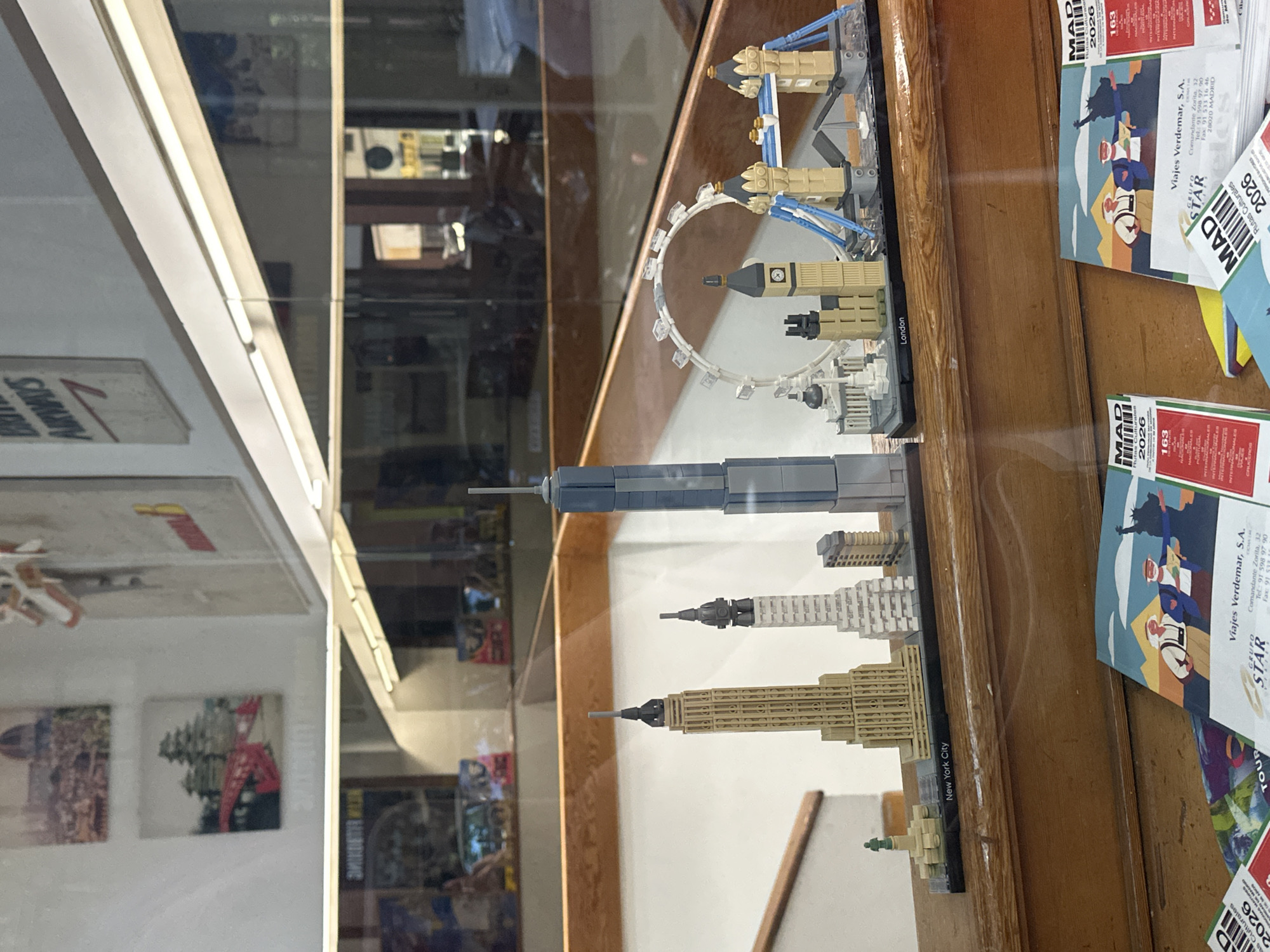 A display case contains Lego models of famous buildings from London and New York City. To the right of the model is a stack of "Viajes Verdemar, S.A." promotional materials which advertise "Rutas Culturales" and list the company address as "Comandante Zorita, 32, 28020 MADRID" (Madrid, Spain). The display case itself seems to be in a shop, with pictures and signage visible in the reflections.