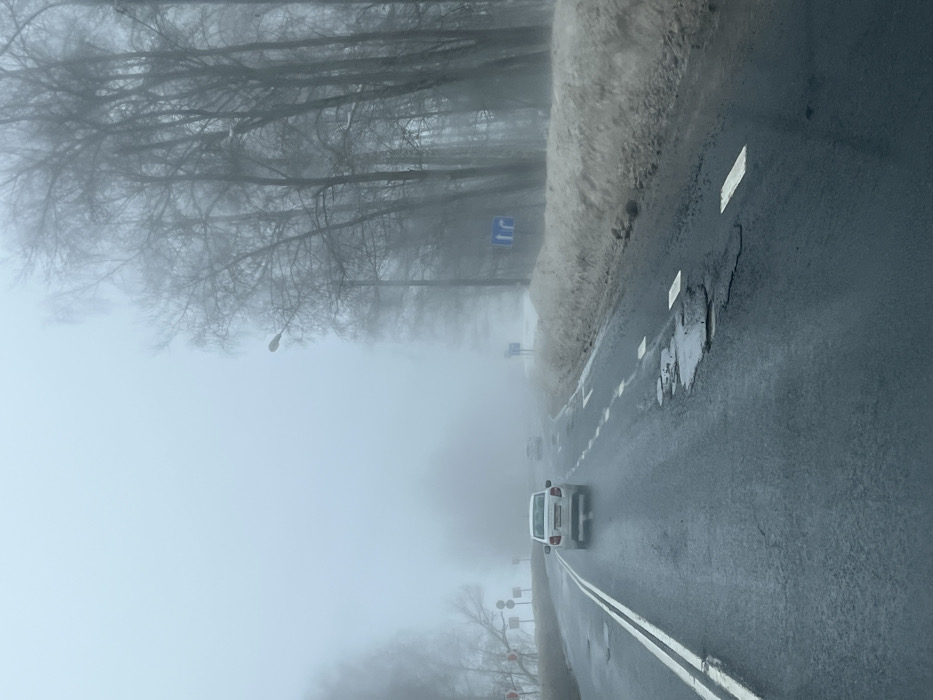 The scene depicts a multi-lane asphalt road in Butyrki, Russia, on a day characterized by dense fog and limited visibility. The road surface is wet and exhibits several potholes, some containing standing water. Lane markings include a double solid white line on the left and broken white lines on the right, delineating lanes.
A white sedan with illuminated taillights is in the foreground, traveling away from the viewer. Its rear license plate appears to be Russian, partially legible as "P796XM 197". Two additional vehicles are faintly visible further ahead on the road, receding into the fog.
To the right of the road, a bank of dirty snow or slush borders the pavement. A line of bare deciduous trees stands further back, partially obscured by the fog. A streetlight on a pole is visible among these trees, and a blue road sign with white arrows indicating a straight path and a right turn is present. Another similar sign is faintly visible further down the road.
The left side of the road shows bare trees and what appear to be utility poles or structures extending into the fog. Distant red and white striped warning barriers or signs are barely discernible through the heavy mist. The sky is uniformly gray and overcast, contributing to the overall hazy and muted light conditions. No people are visible.