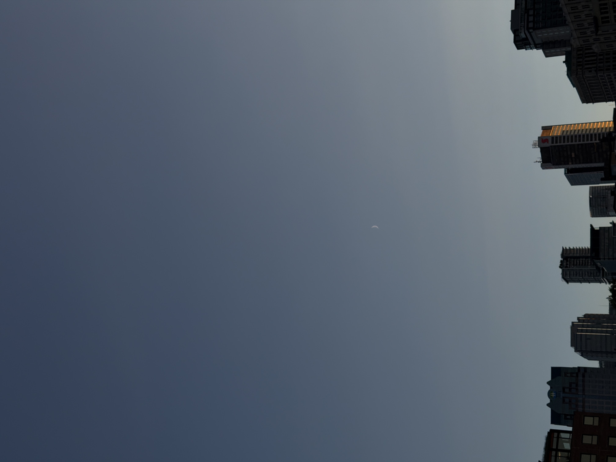 A crescent moon is visible in a clear, blue sky above a cityscape. The buildings in the foreground appear to be part of the downtown core of Vancouver, Canada, with several modern high-rise structures and some older, lower buildings. The sun is not visible, but the light suggests it may be late afternoon or early morning. There are no people or animals visible in the image. The overall impression is one of an urban environment under a vast, open sky.
