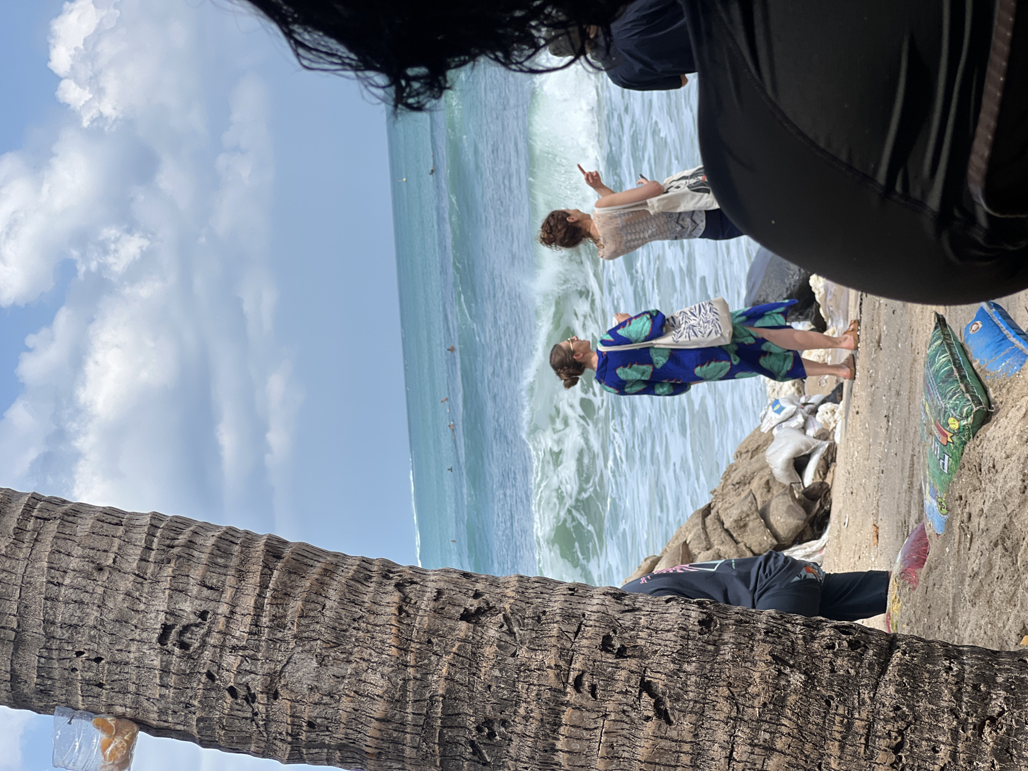 The image depicts a scene on a beach, likely in Kuta, Indonesia, with a focus on the interaction of people with the ocean and the surrounding environment. In the foreground, a large palm tree trunk occupies the left side, its rough bark texture prominent. A clear plastic container with pieces of orange fruit is affixed to the tree.
Further into the scene, several individuals are visible. Two women stand near the water's edge, one wearing a blue and green patterned dress and the other a patterned sleeveless top. The woman in the patterned top is pointing towards the ocean. Another person, mostly obscured by the foreground, appears to be standing to the right. In the distance, several surfers are seen in the turquoise ocean, with waves breaking closer to the shore. The beach itself appears somewhat littered, with various objects including bags and debris scattered on the sand, some partially covered by sand. The sky is cloudy with patches of blue. The overall impression is a busy beach scene with a mix of recreational activity and the presence of environmental elements.