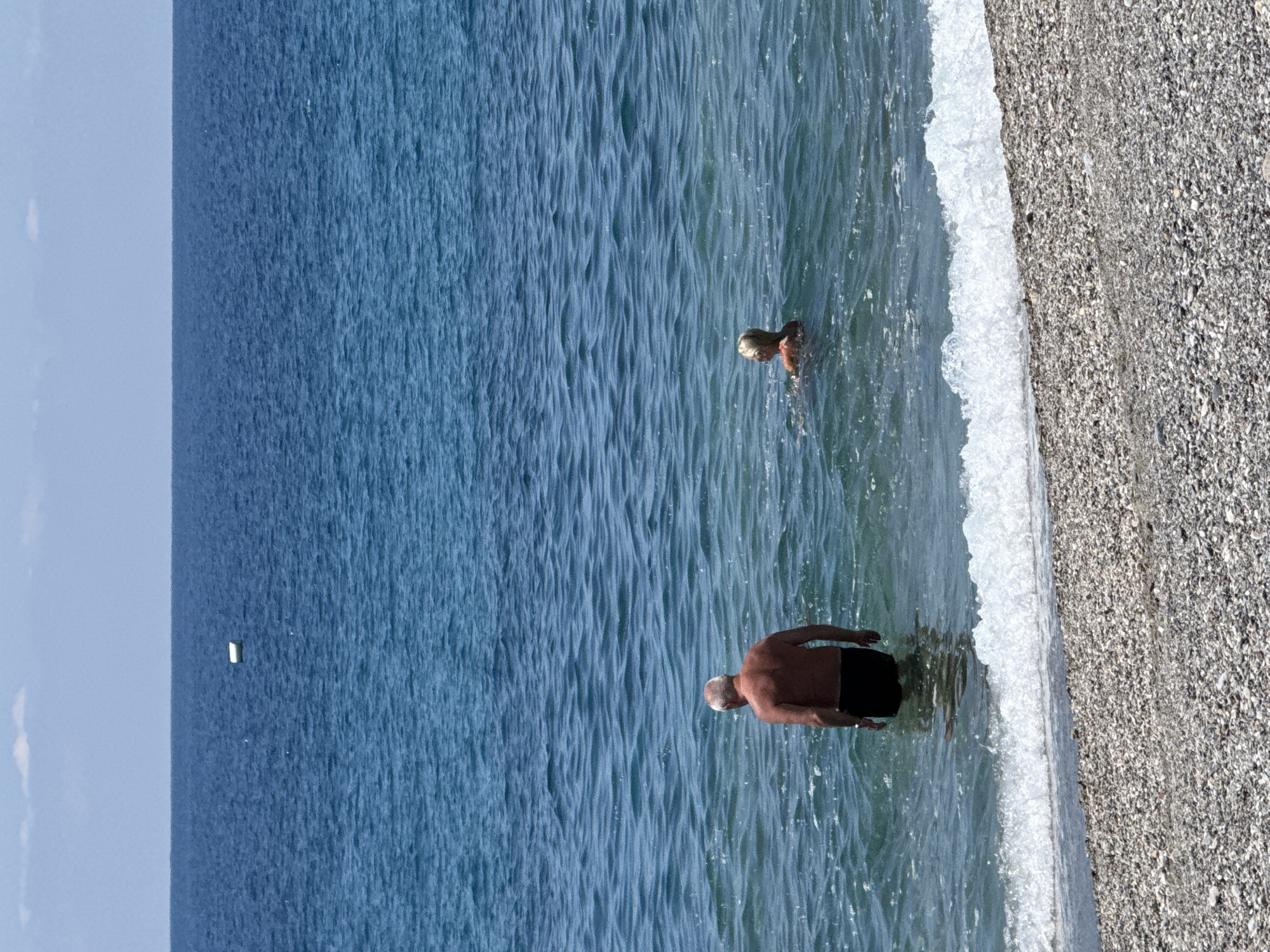 The image displays a serene beach scene under bright daylight. The natural orientation, when rotated counter-clockwise, shows a clear blue sky at the top, meeting a vast expanse of blue-green sea at the horizon. The sea occupies the majority of the frame, gradually transitioning to a pebbly shore at the bottom right.
Two individuals are present in the water. Closer to the shore, an older man with light or balding hair is standing waist-deep in the water, facing away from the viewer and slightly to the right. He is shirtless and wearing dark swim trunks. Further out in the sea, a second person, appearing smaller with light-colored hair, has their head and shoulders visible above the water, seemingly swimming or wading.
A white, foamy wave breaks gently along the edge of the pebbly shore, just behind the man. The water exhibits subtle ripples and reflections. In the far distance, on the horizon line where the sky meets the sea, a small, white, rectangular object is visible, possibly a distant boat or a buoy. The overall atmosphere suggests a calm, sunny day at a beach. No visible text is present.