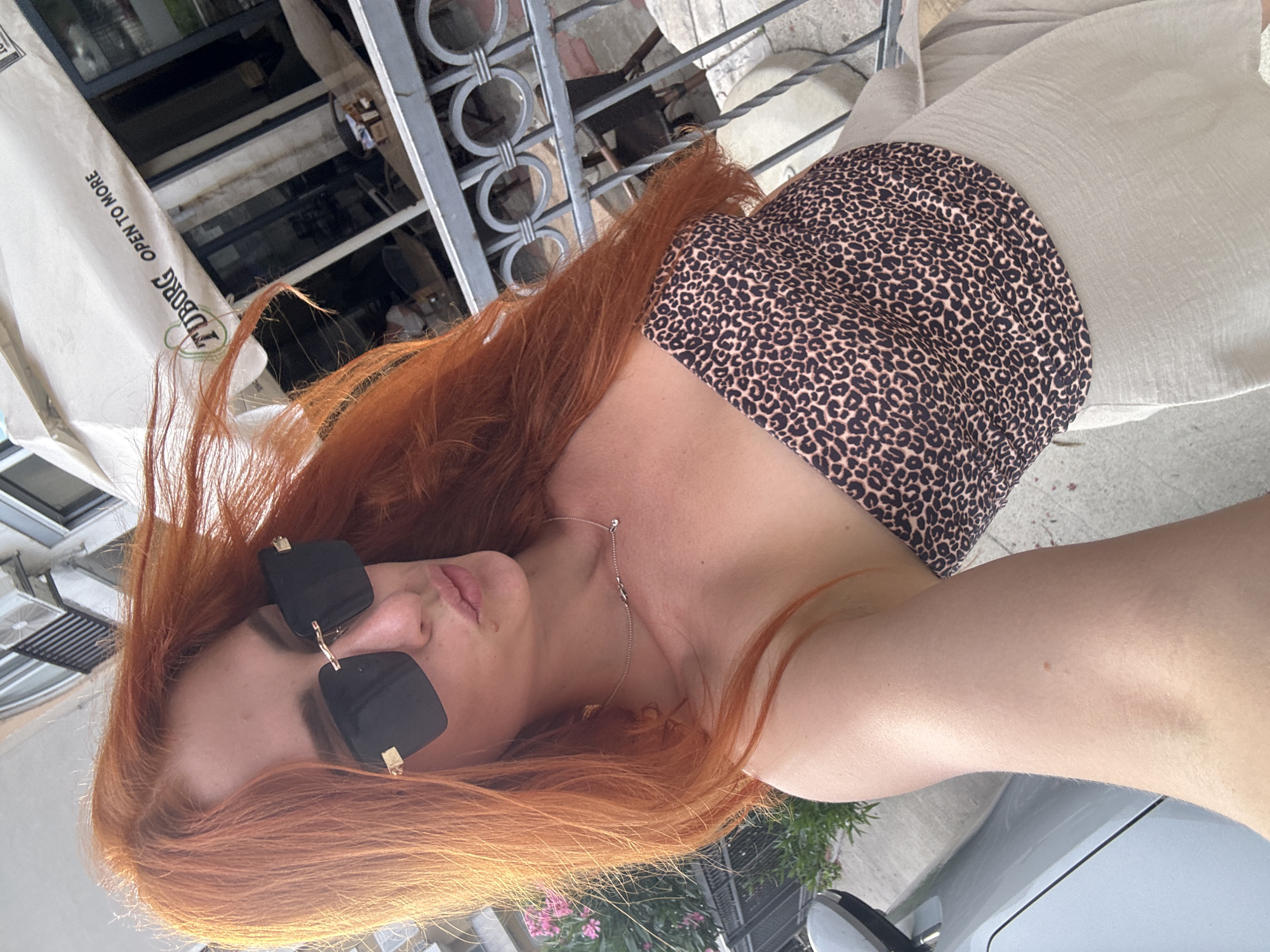 The image shows a woman with long, vibrant red hair and fair skin. She is wearing large, black, square-shaped sunglasses with gold accents and a leopard-print bikini top. A delicate silver necklace is visible around her neck. She is outdoors, with a light-colored, textured fabric draped over her shoulder and a hint of a car in the lower right corner.
In the background, there are hints of an urban or resort setting. A metal railing with decorative circular patterns is visible behind her, suggesting a balcony, terrace, or fence. Beyond the railing, there appears to be a building with windows and some greenery with pink flowers. A portion of a white umbrella with dark text is also visible on the left side of the image.
The lighting suggests it is daytime, likely sunny, as indicated by the bright illumination and the presence of sunglasses. The overall mood appears casual and relaxed, possibly during a vacation or a leisurely outing.
Visible text on the umbrella reads "JJUD.BORG OPEN TO MORE". The location context is Budva, Montenegro.