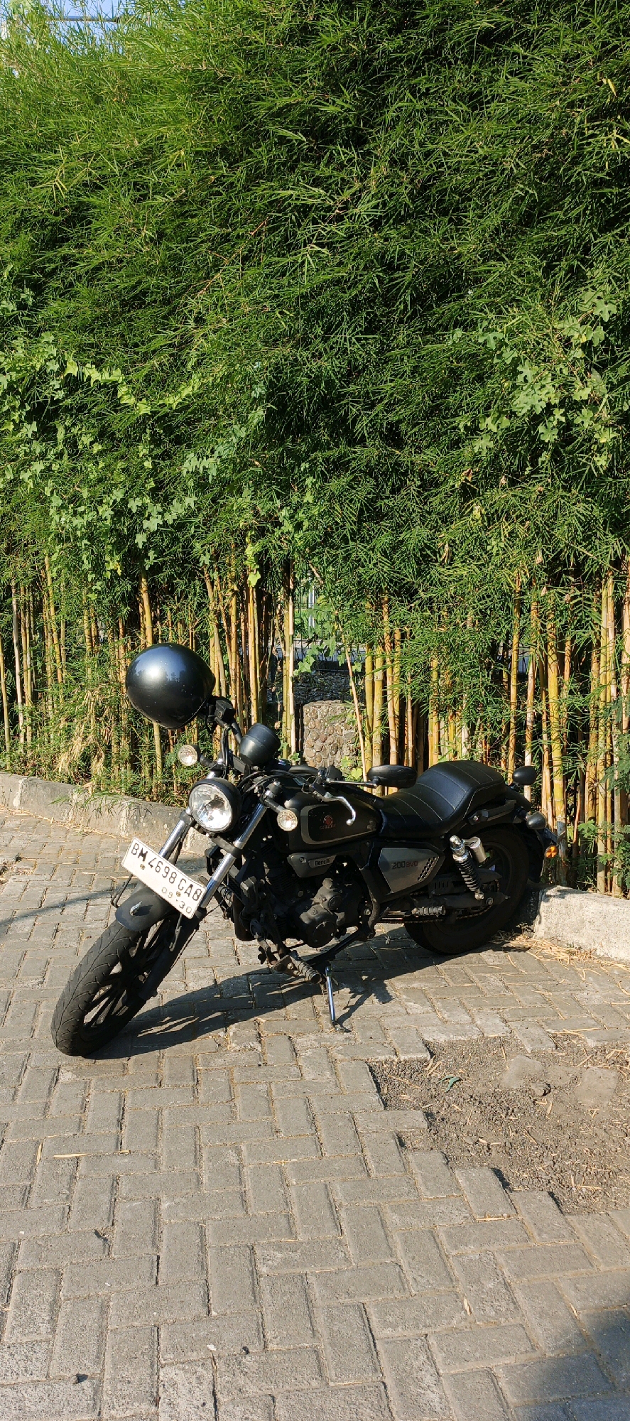 A black motorcycle is parked on a brick paver pathway in front of a dense bamboo grove. The motorcycle is oriented facing left, with its front wheel slightly turned. A dark, spherical helmet is attached to the handlebars. The license plate on the front of the motorcycle is visible and reads "BM 4698 GA8". The bamboo stalks are yellow-green, and the leaves are a vibrant green. Sunlight casts a shadow of the motorcycle on the pavers. The environment appears to be outdoors, possibly in a park or a roadside area in Trihanggo, Indonesia, given the commonality of bamboo groves and paver pathways in such locations. There are no people visible in the image.