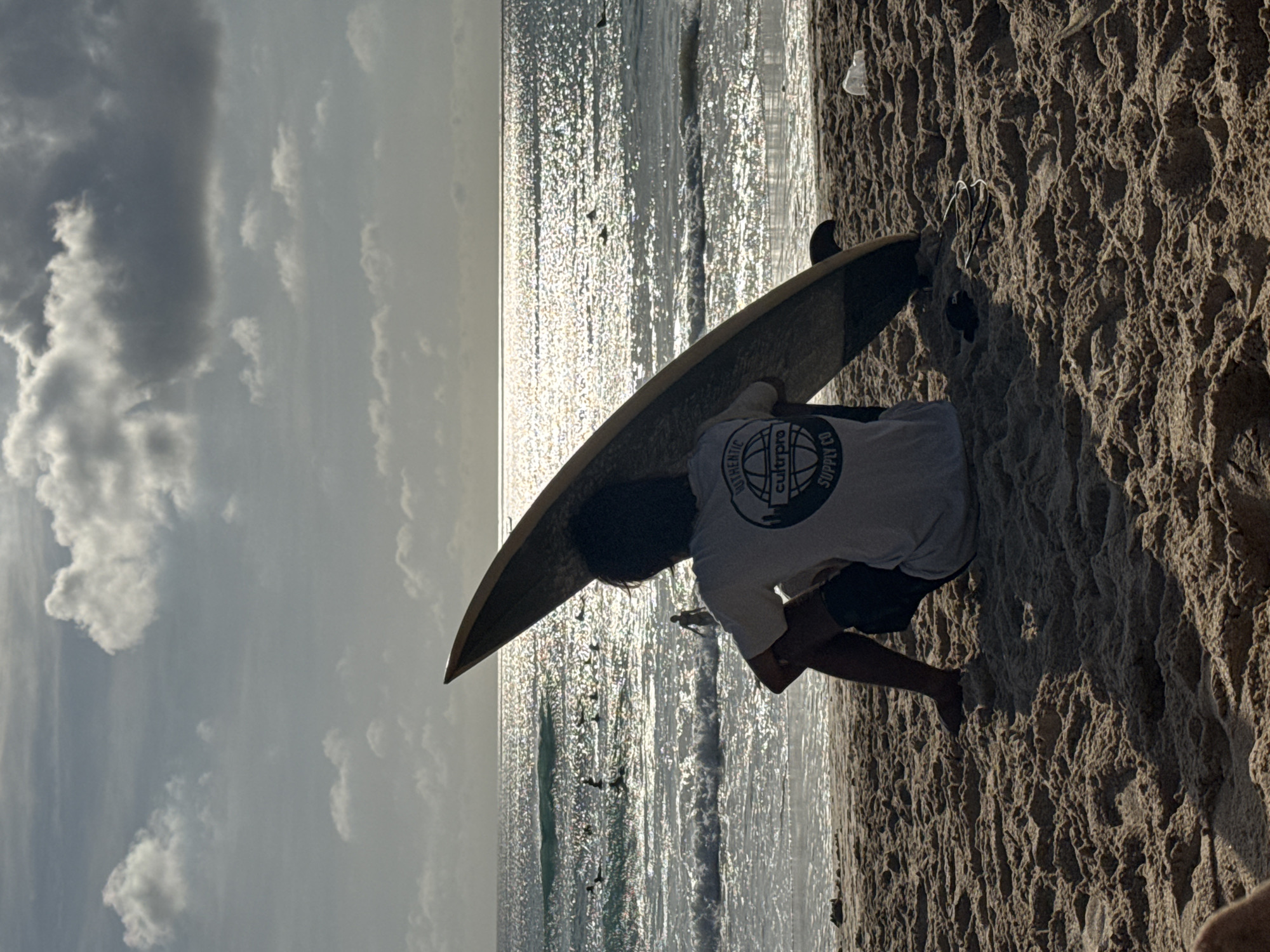 A man is kneeling on the sand next to a surfboard at the beach in Kuta Selatan, Indonesia. He is wearing a white t-shirt with black shorts. The sea is behind him and to the left. The sky is cloudy.