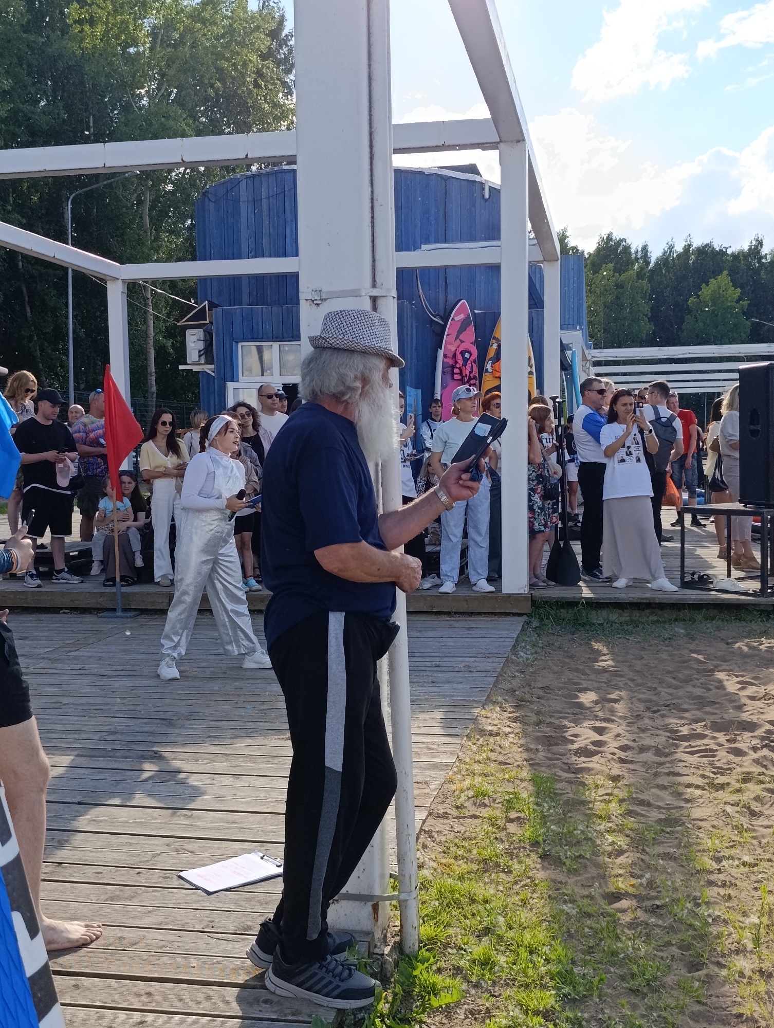 A group of people are gathered on a wooden deck near a sandy area, likely a beach or waterfront, in Chaikovsky, Russia. In the foreground, an older man with a long white beard and a patterned hat is speaking into a microphone while holding a tablet. He is wearing a dark blue t-shirt and black pants with a grey stripe. Behind him, a young woman in a silver outfit is also speaking into a microphone. She is standing in front of a group of spectators. Several surfboards are visible leaning against a blue building in the background. The environment suggests an outdoor event, possibly a competition or festival, taking place on a sunny day.