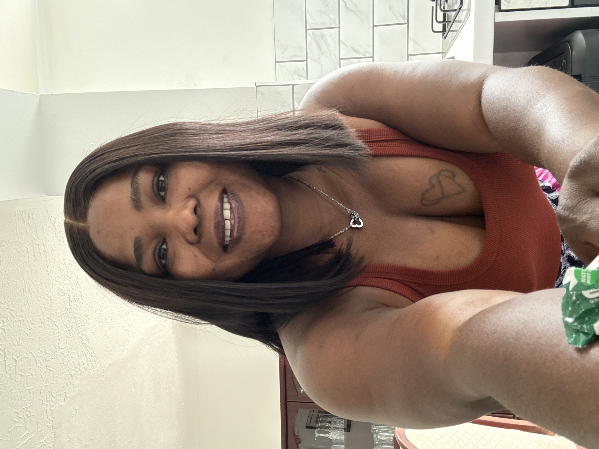 A medium shot captures a Black woman with dark hair and dark lipstick, smiling at the camera. She is wearing a rust-colored tank top and a silver necklace with a heart pendant. A tattoo is visible on her chest. The background includes kitchen cabinets with glassware, a tiled backsplash, and a stovetop. The overall scene suggests a home environment, possibly a kitchen, in Darlington, United Kingdom.