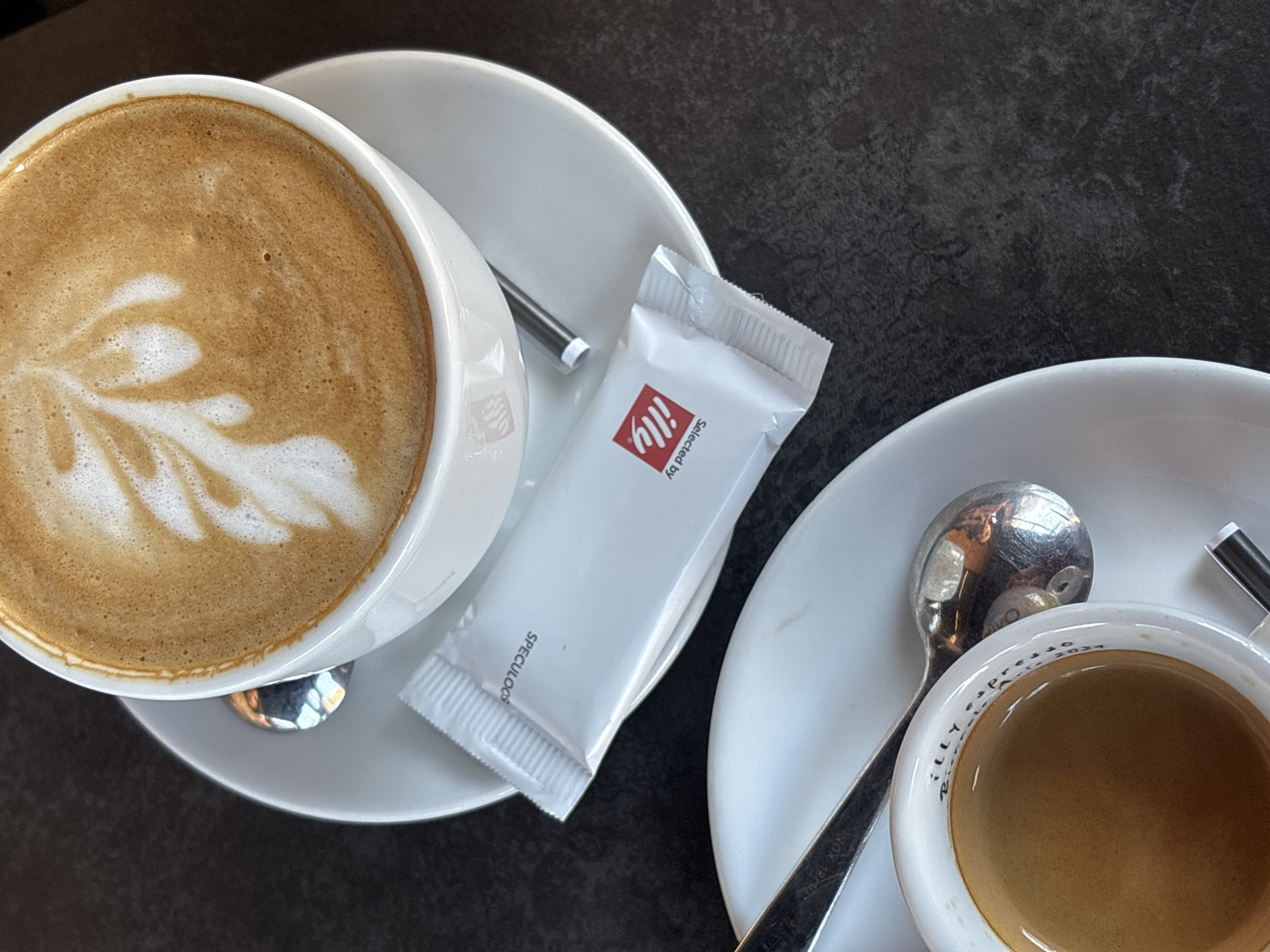 The image captures an overhead, close-up view of two coffee servings arranged diagonally on a dark, textured tabletop within an indoor cafe environment in Varna, Bulgaria. In the upper portion, a white ceramic cup contains a light brown beverage, likely a cappuccino or latte, adorned with white foam art depicting a stylized leaf or fern pattern. This cup rests on a white ceramic saucer, which also holds a silver-colored spoon and a sealed white rectangular packet. The packet is branded with 'Selected by Illy' in red lettering on one side and 'SPECULOOS' in black lettering on the other. In the lower portion of the frame, partially visible, is another white ceramic cup, rimmed with 'ILLY espresso' branding, containing a darker brown beverage, presumably an espresso. This cup is also positioned on a white ceramic saucer with a silver-colored spoon. No individuals are visible, and no active interactions are depicted; the scene presents a static arrangement of beverages and associated items.