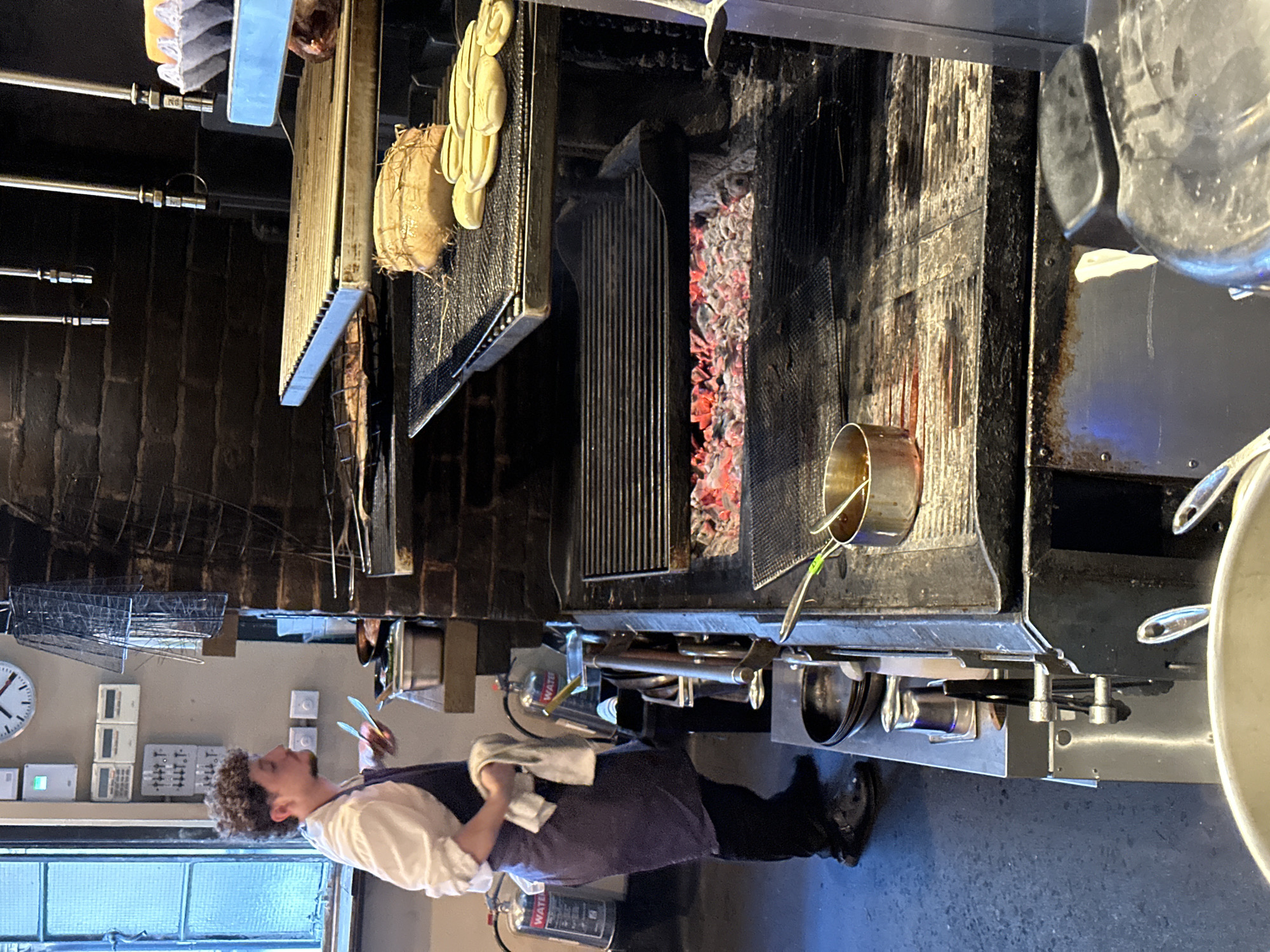 The image depicts the interior of a commercial kitchen, likely a restaurant. A man dressed in a white shirt and dark apron stands near a large grill with glowing red embers. He appears to be a chef, holding utensils. Above the grill, various cooking utensils and equipment are hanging, including pans and tools. The kitchen environment includes brick walls, stainless steel surfaces, and typical restaurant fixtures like a clock and switches. The location is suggested as London, United Kingdom. The overall impression is of a busy, functional kitchen environment focused on food preparation.
