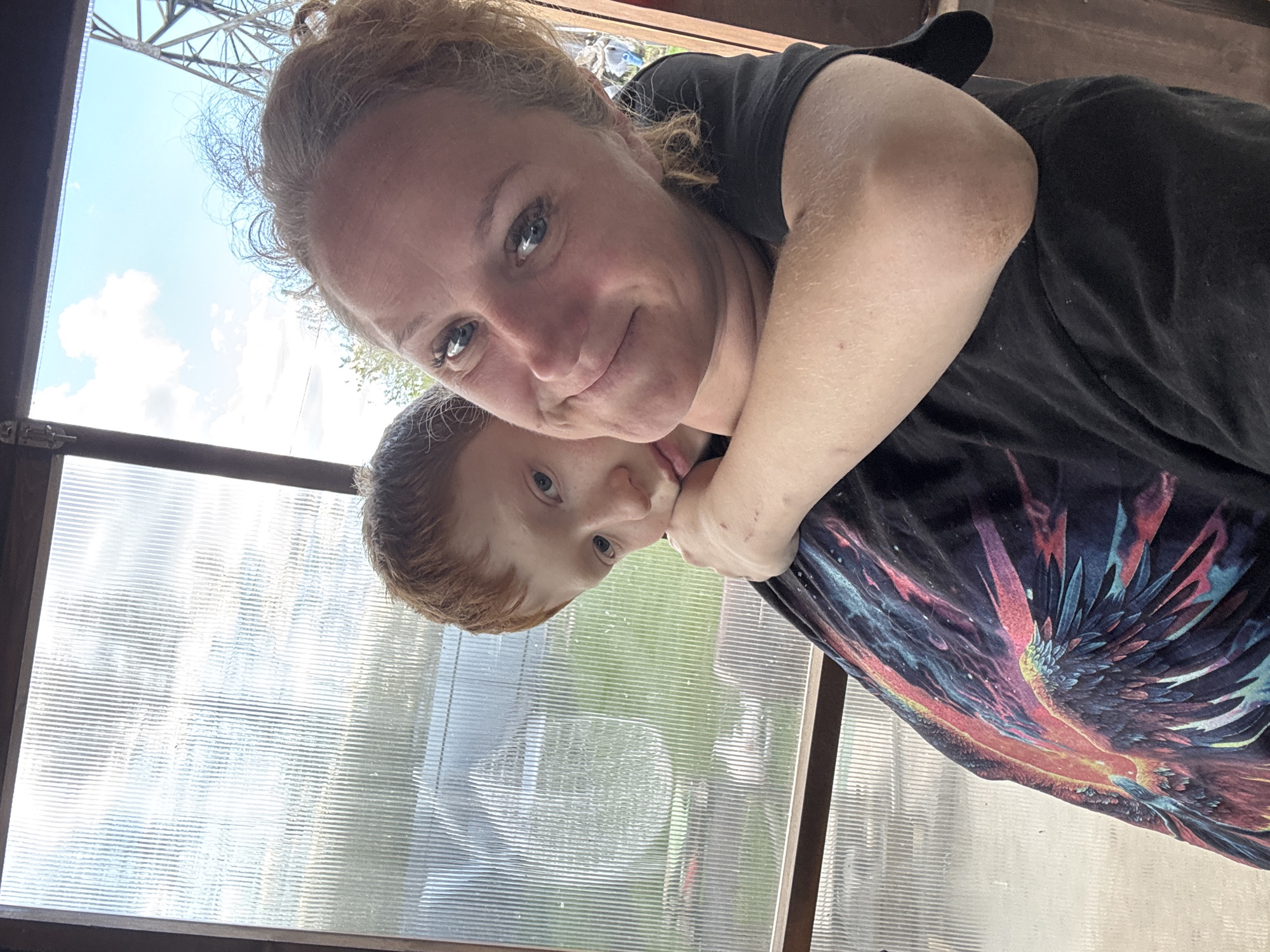 The image is a selfie of a woman and a child, likely a mother and son. The woman has fair skin, reddish-blonde hair pulled back, and blue eyes. She is wearing a dark-colored t-shirt. The child has red hair, fair skin, and blue eyes. He is leaning on the woman, possibly being hugged. The woman's arm is around the child.
Behind them is a window with a view of trees and sky. The window appears to be made of translucent ribbed plastic or glass, possibly part of a greenhouse or enclosed porch. The lighting suggests it is daytime.
Without further context, it is not possible to verify if this photo was taken in Chaikovsky, Russia or provide more information.