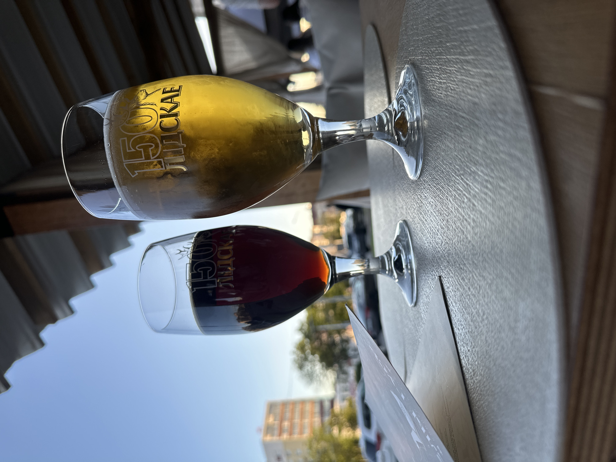 Two glasses of beer sit on a wooden table, one filled with a pale golden lager, the other with a dark, rich stout. The glasses are etched with the text "150 ЛЕТ СКИ". The scene appears to be outdoors, with a blue sky visible in the background, suggesting it's a sunny day. A hint of a building and trees indicates an urban setting, possibly a cafe or restaurant. The lighting suggests it's late afternoon or early evening, with the sun casting long shadows. The overall mood is relaxed and casual. The image is oriented vertically, as if the camera was held at an unusual angle. The context provided places the scene in Minsk, Belarus.
