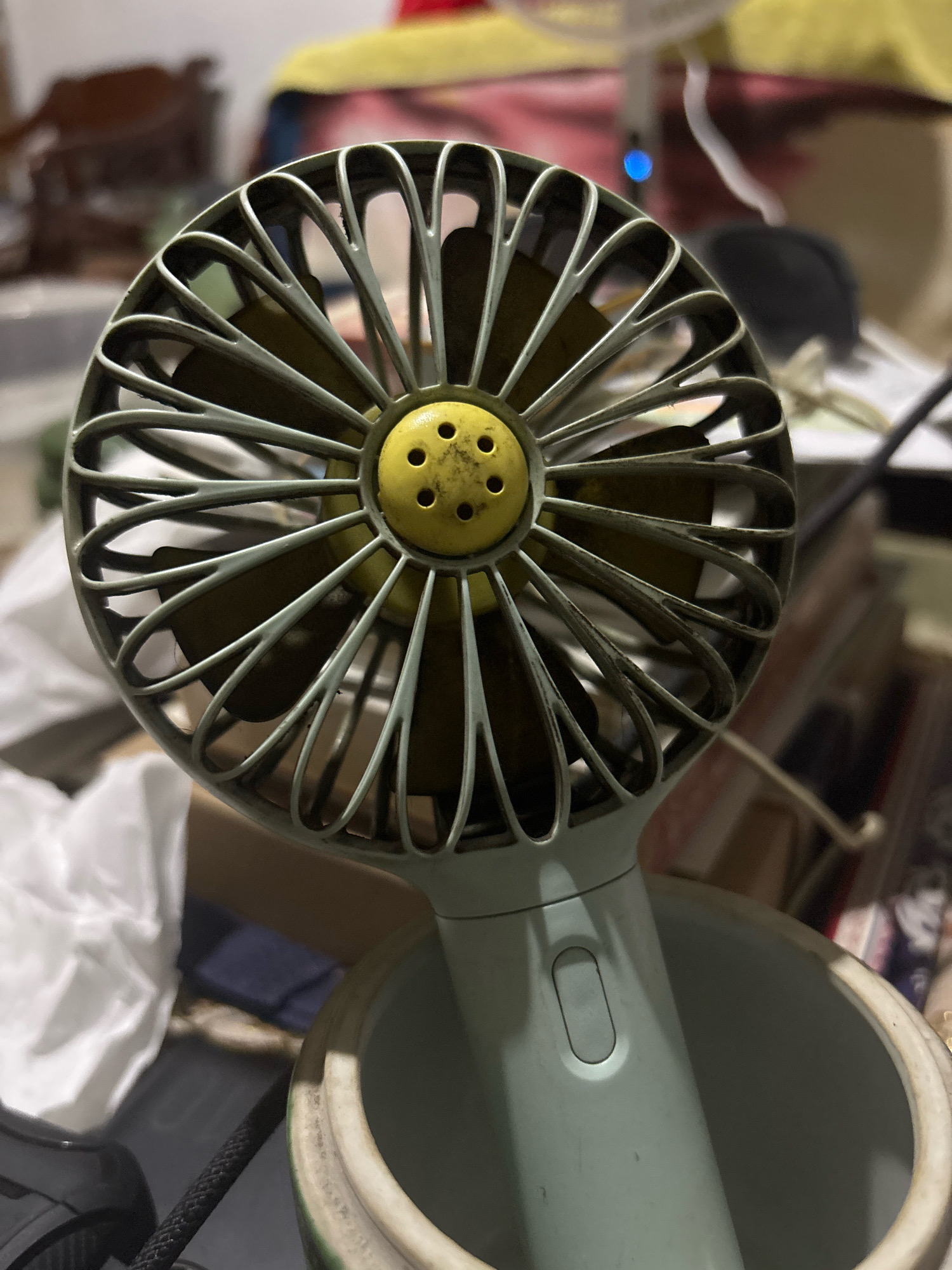A close-up shot shows a light blue portable fan with a yellow central hub featuring six small holes. The fan's blades are visible through its protective grille, which is also light blue. The fan is placed inside a cream-colored ceramic container with a greenish-white rim. In the background, various objects are blurred, including what appears to be furniture, papers, and fabric, suggesting an indoor setting. The overall scene is cluttered and appears to be a home or office environment. There is no indication of the city of Bekasi, Indonesia in the image.