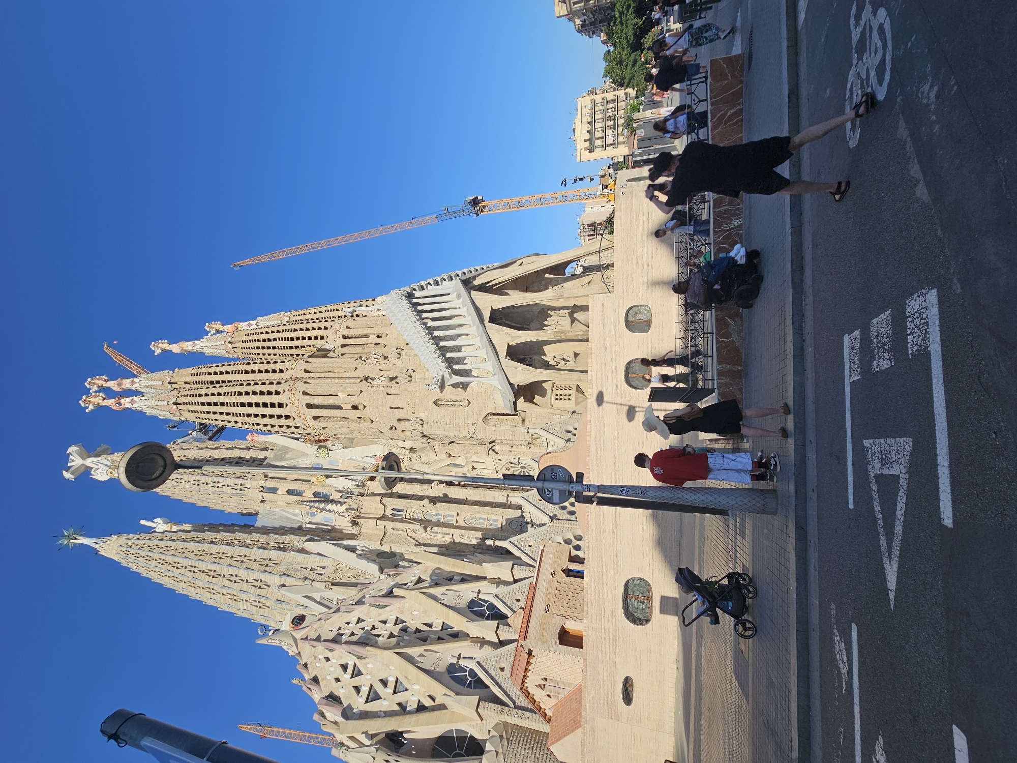 This media file depicts a sunny day in Barcelona, Spain, with the iconic Sagrada Familia basilica as the central focus. The basilica, still under construction, features its characteristic intricate, organic architectural style. Cranes are visible, indicating ongoing building work.
Several people are present in the scene. On the right side of the image, a man in black shorts and a black t-shirt walks on a sidewalk adjacent to a street. Another person in a red shirt and white shorts stands near a stroller. Other individuals can be seen in the background, some walking along the sidewalk and others gathered near the basilica.
The sky is a clear, vibrant blue, suggesting good weather and likely midday or late afternoon given the shadows. The ground is paved with a mix of sidewalk tiles and asphalt, marked with white lines indicating traffic lanes and a bicycle symbol. The overall atmosphere appears relaxed, with people enjoying the exterior of the architectural marvel. No legible text is visible in the image.