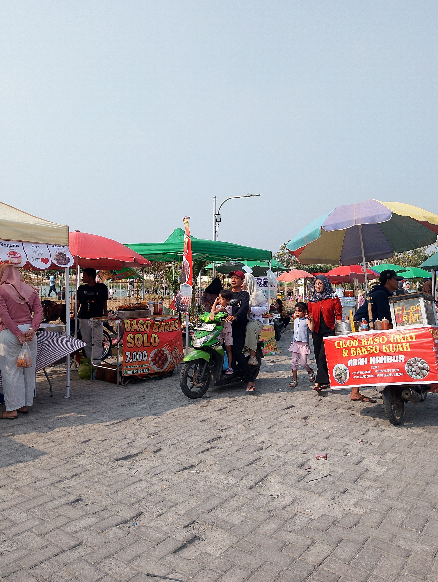 The image depicts a street food market scene in Karawang, Indonesia, on a bright, clear day. The foreground is paved with grey brickwork, showing some wear and tear. Several food stalls are set up, identifiable by their colorful umbrellas, some striped in yellow, purple, and blue, and others solid red or green.
In the center, a green motorbike is ridden by a man wearing a red cap and a black t-shirt. A woman in a white headscarf and black shirt sits behind him, and two young girls are with them. The man is looking ahead, while the woman appears to be looking back.
To the left, a woman in a pink hijab and grey pants stands with her back to the camera, holding a plastic bag containing snacks. Next to her is a food stall with a checkered tablecloth and a sign that reads "BASO BAKSO SOLO 7.000". A vendor in a black t-shirt is visible behind the stall, and a gas tank is placed on the ground.
To the right, another food cart is prominent, with a large red banner that says "CILOK BASO URAT & BASO KUAH ABAH MANSUR". The banner also lists the available food items. A woman in a red jacket and a grey headscarf is walking alongside a young girl in a pink dress. A man with a black cap stands near the cart, seemingly attending to customers.
In the background, more food stalls and people can be seen, suggesting a lively market atmosphere. A metal fence separates the market area from a body of water, likely a river or lake, where more people are visible. Overhead, a street light is mounted on a pole. The sky is a pale blue, indicating good weather.
