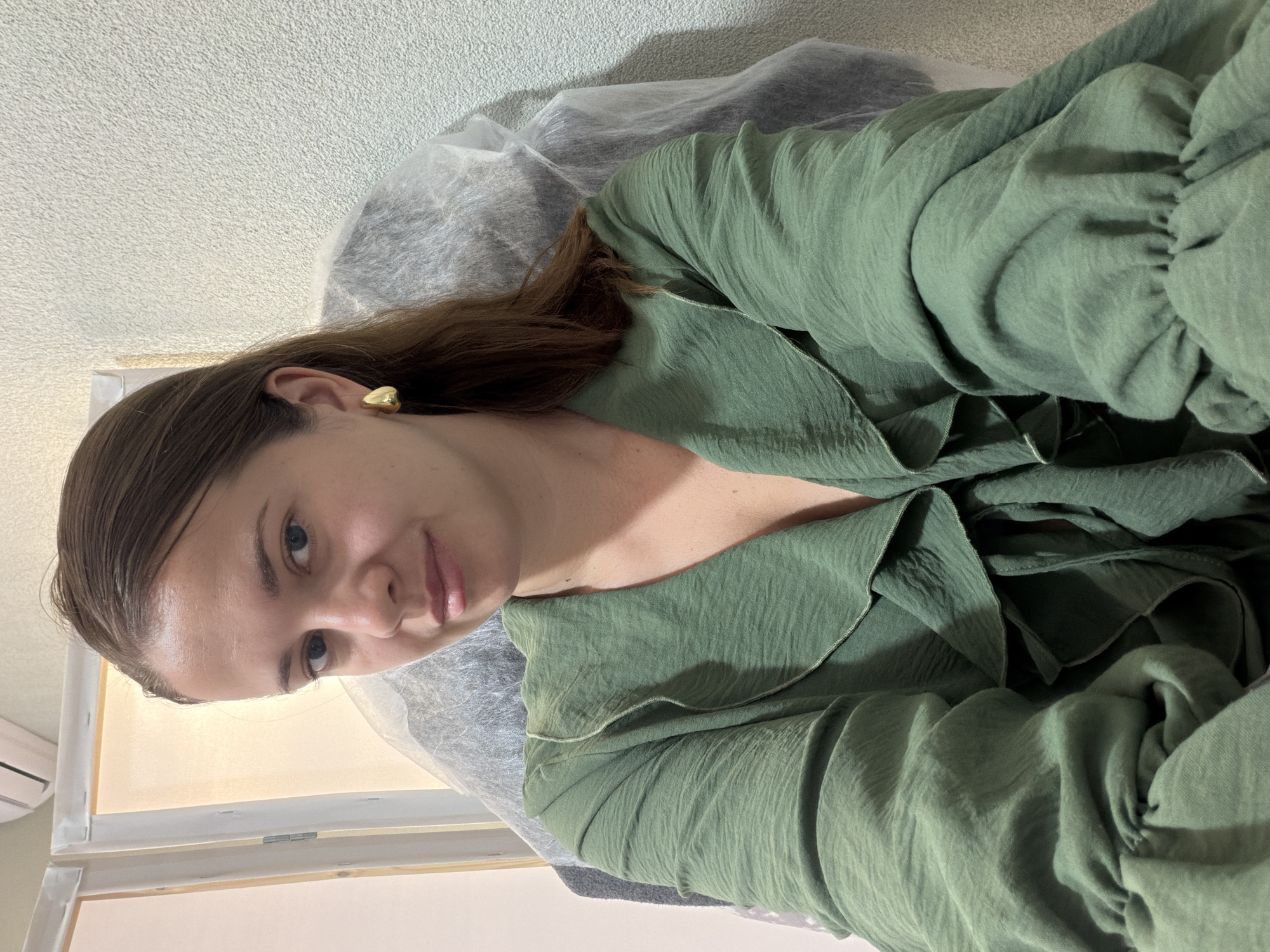 A young woman with long brown hair and blue eyes is seated indoors, looking directly at the camera. She is wearing a green ruffled blouse and a large gold earring on her right ear. The background consists of a light-colored textured wall and what appears to be a white screen or canvas. There is also a clear plastic covering draped behind her, obscuring a portion of the background. The lighting is soft and appears to be artificial. The image does not contain any specific visual cues that would indicate its location within the city of Sochi, Russia.