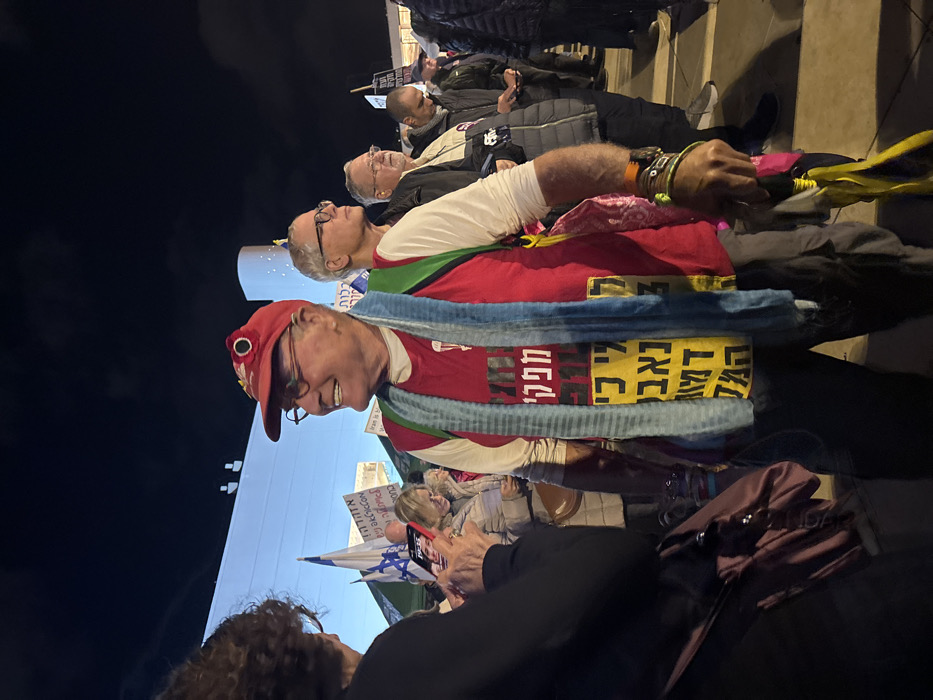 A nighttime outdoor scene captures several individuals. An older man with glasses and a red cap smiles, wearing a red t-shirt and a blue scarf. Text in Hebrew is visible on his clothing. Behind him, other people stand or sit, some looking upwards. An Israeli flag is partially visible, and a woman in the foreground appears to be holding a phone. A building with lights is in the background.