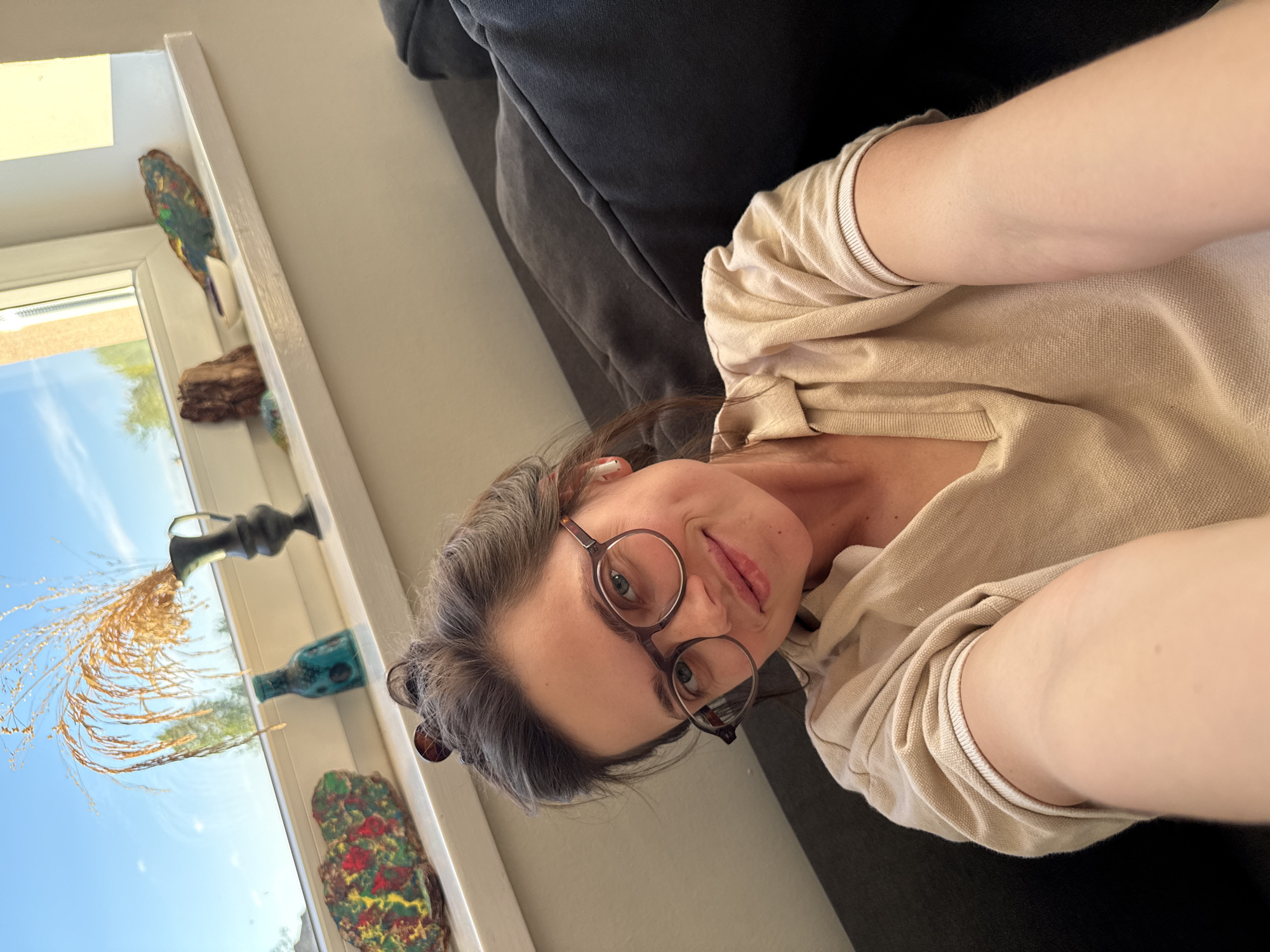 A woman is captured in a selfie, taken indoors, presumably in a home setting. She is reclining on a dark grey sofa or cushion, with the image rotated 90 degrees clockwise. She has dark hair with visible grey strands, wears round, dark-rimmed glasses, and a light-colored (beige or cream) polo-style shirt. Her face is turned towards the camera, showing a subtle, relaxed smile, and she appears to be wearing white wireless earbuds.
Behind her, a white window frame reveals a bright, clear blue sky with some distant green foliage, indicating a sunny daytime. On a narrow shelf or window sill beneath the window, several decorative items are arranged. These include a colorful, abstract sculpture resembling a bird, a piece of natural wood, an ornate dark-colored vase holding dried golden grass or reeds, a teal or turquoise bottle, and another vibrant, abstract decorative piece at the bottom. The overall impression is one of a casual, bright, and personalized indoor space. There is no visible text in the image.