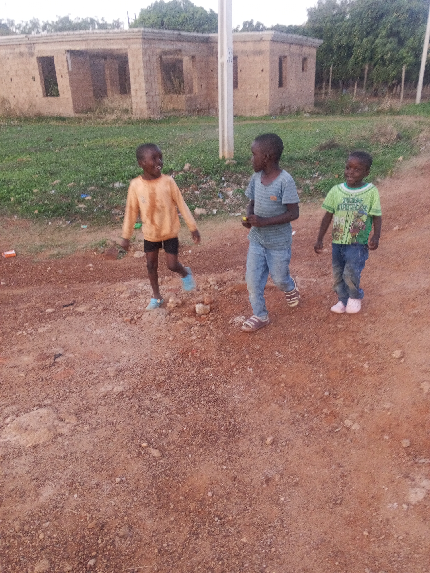 Three children are walking in a dirt area. The child on the left wears a yellow shirt and black shorts. The child in the middle wears a blue striped shirt and jeans. The child on the right wears a green shirt with the word "Turtles" on it and jeans. Behind the children is a partially constructed building and green vegetation. The location is in Bukuru, Nigeria.