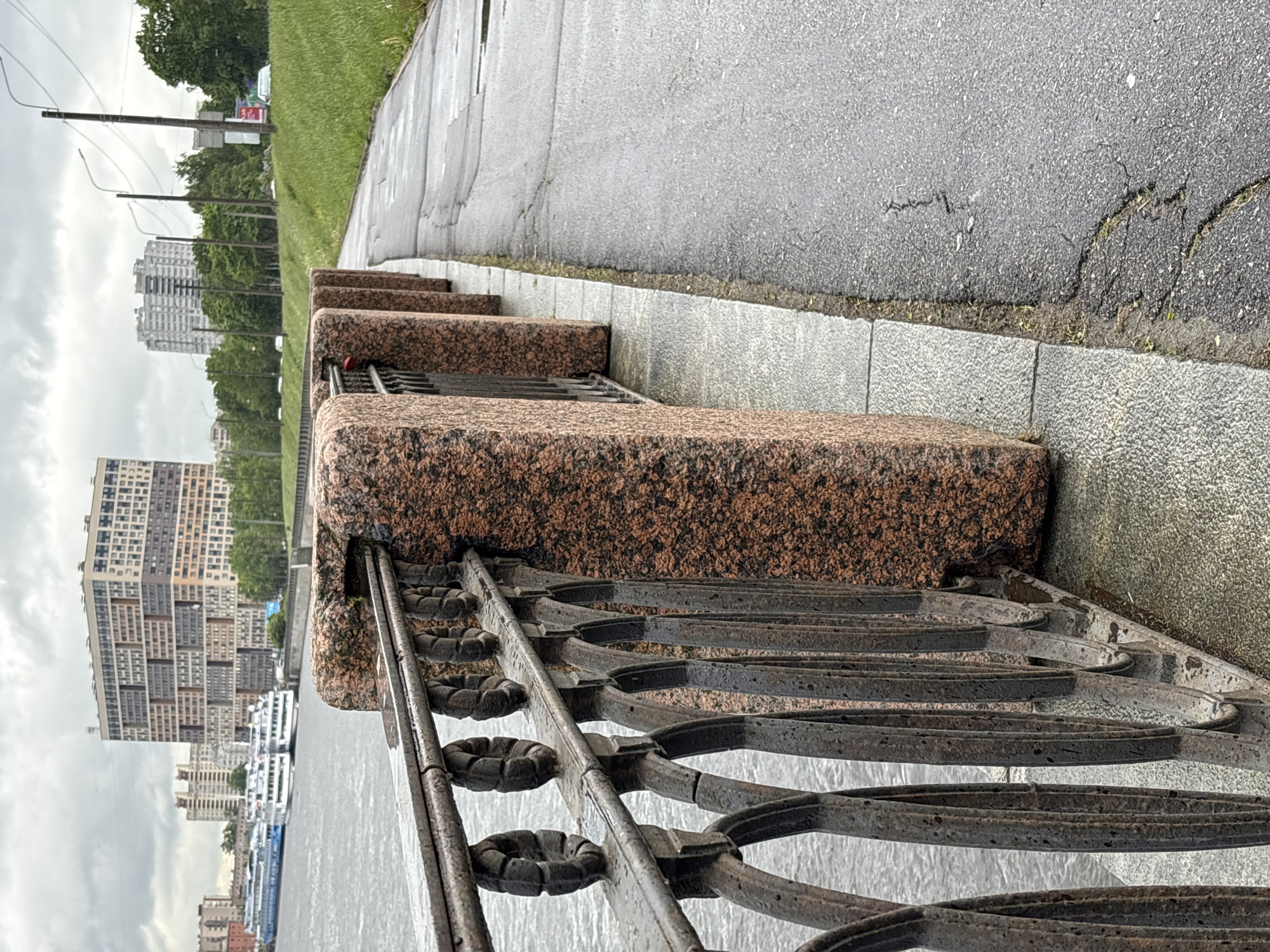 The image shows a section of the granite embankment in Saint Petersburg, Russia. The foreground features a decorative iron railing attached to rust-colored granite blocks. Behind the railing, water can be seen. A gray sidewalk runs parallel to the embankment, followed by a strip of green grass and trees. In the background, several tall buildings of the city rise. The weather appears overcast and damp.