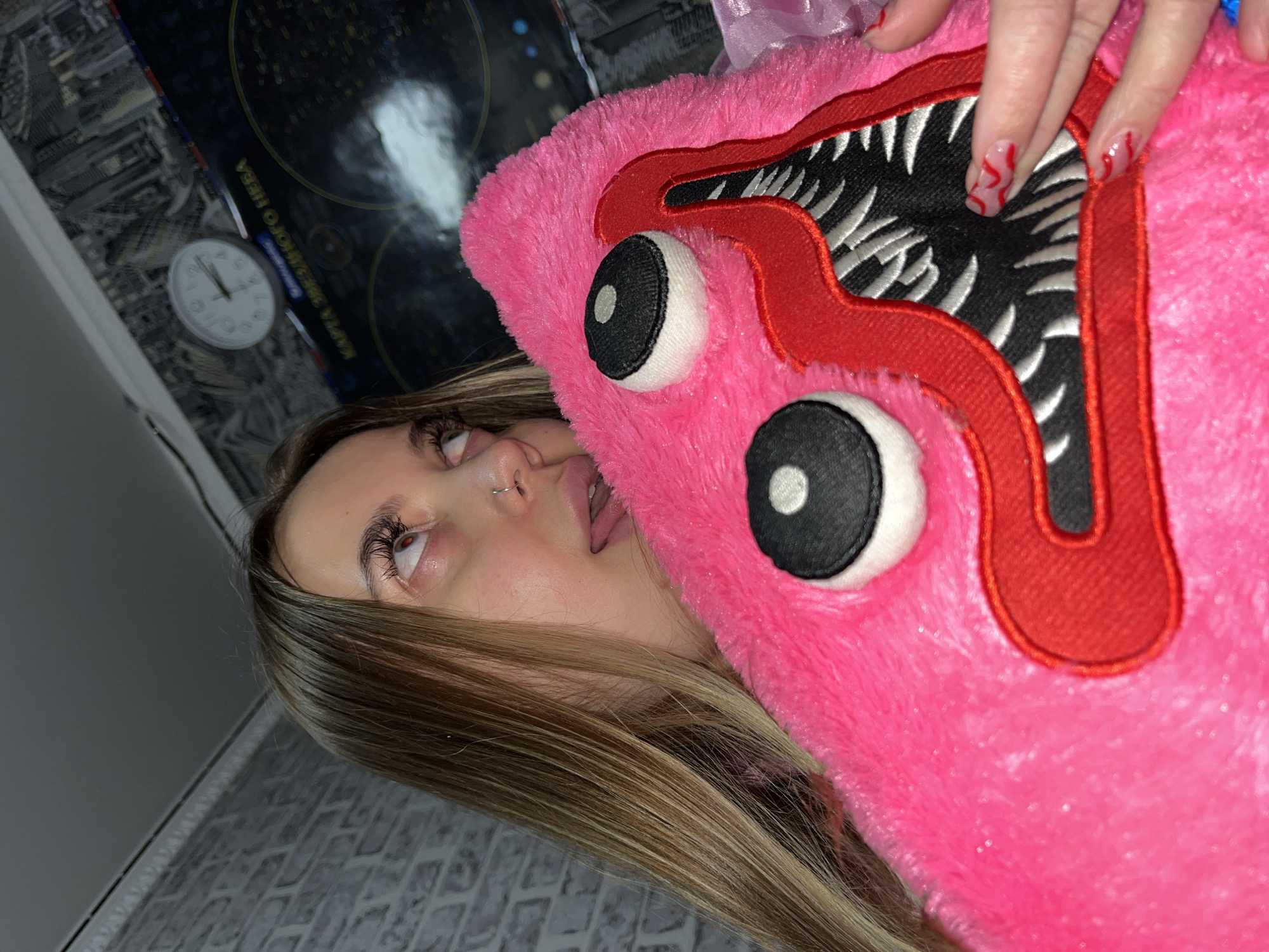A woman with long brown hair and a nose piercing is lying on a surface with patterned wallpaper. She's holding a pink plush toy with large, cartoonish eyes and a red, stitched mouth filled with white teeth. The toy is partially obscuring her face. A clock on the wall reads approximately 11:23, and a poster is also visible in the background. Based on the description of the location as Novopokrovskaya, Russia, the image was likely taken in an indoor setting, possibly a residential home.
