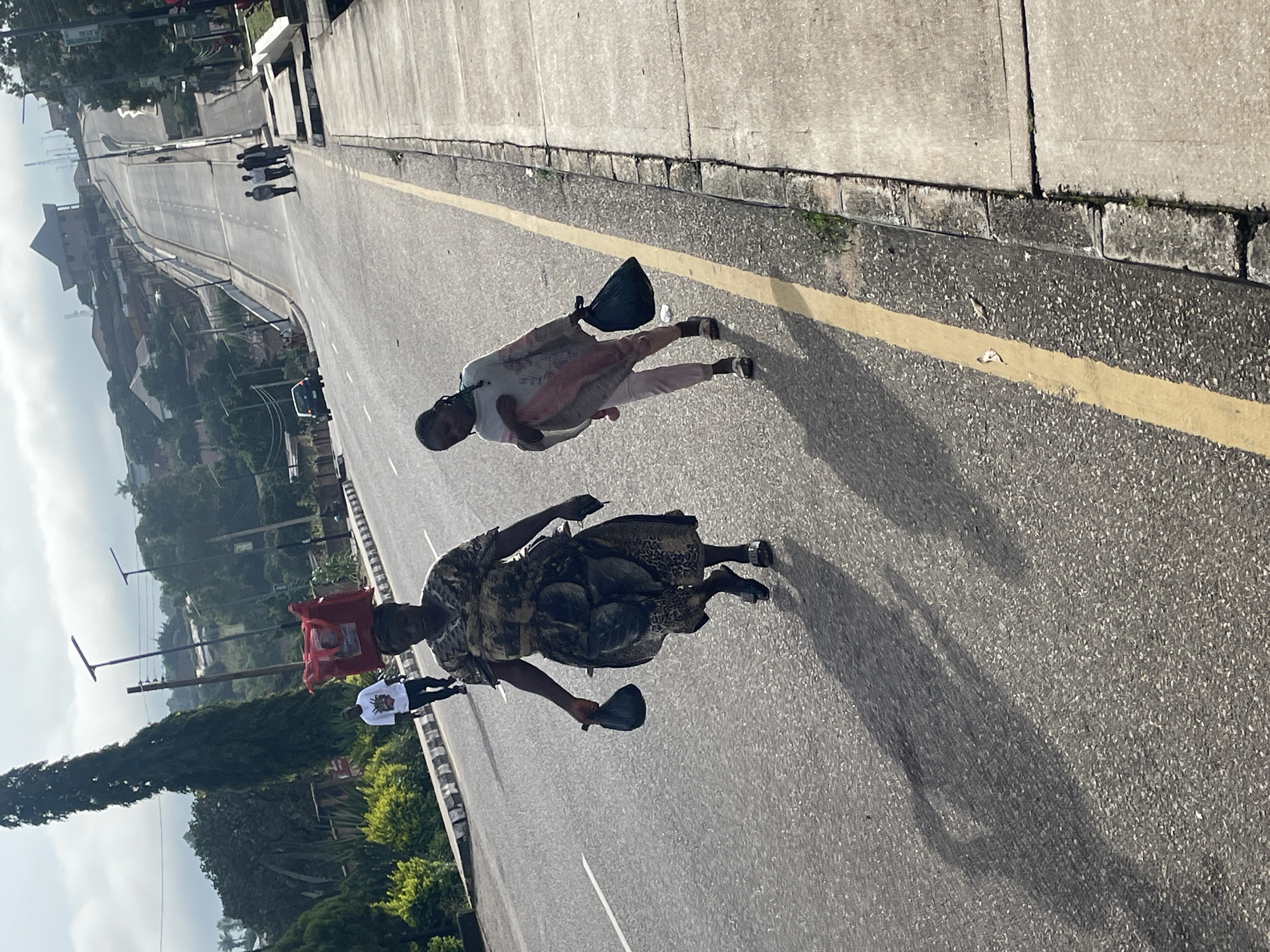 The image captures a sunny day in the city of Jos, Nigeria, with a street scene unfolding. A woman, dressed in a patterned dress and holding a dark bag in her left hand, walks on the asphalt road. A young girl, in light-colored pants and a light grey garment, walks beside her, carrying a dark bag in her right hand and a light pink bundle in her left. They are both casting long shadows on the road, indicating the low angle of the sun. In the background, a man in a white t-shirt and dark pants walks carrying a red bag on his head. Further up the sloping road, a group of men are walking together, and a car is visible on the left side of the road. The environment suggests a residential or commercial area with buildings and lush greenery lining the street.