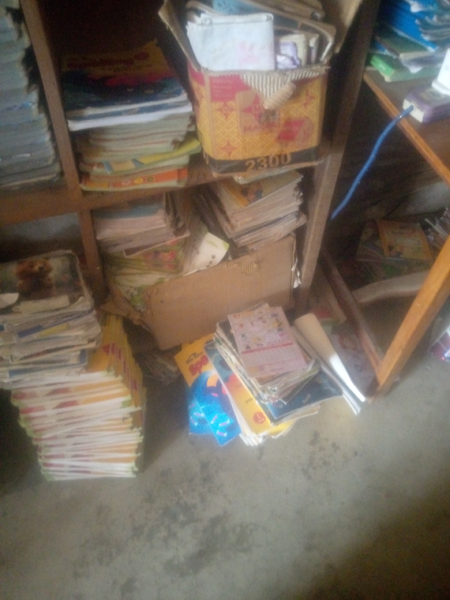 The image depicts a cluttered room filled with books and papers. A wooden shelf unit is visible, with books stacked on multiple shelves and on the floor. In the upper left corner, a stack of books with blue and grey covers is seen. To the right of this stack, on the same shelf, are more books, some with colorful covers. A cardboard box filled with paper items sits on the middle shelf, with the number "2300" visible on its front. To the right of the box, a blue cord trails across the shelf. The lower shelves and floor are also covered with stacks of books and papers, some of which appear to be children's books with cartoonish illustrations. A brown cardboard box is placed on the floor near the bottom center. The overall impression is one of disarray and abundance of reading material. The environment appears to be indoors, possibly a storage area or a room dedicated to books.