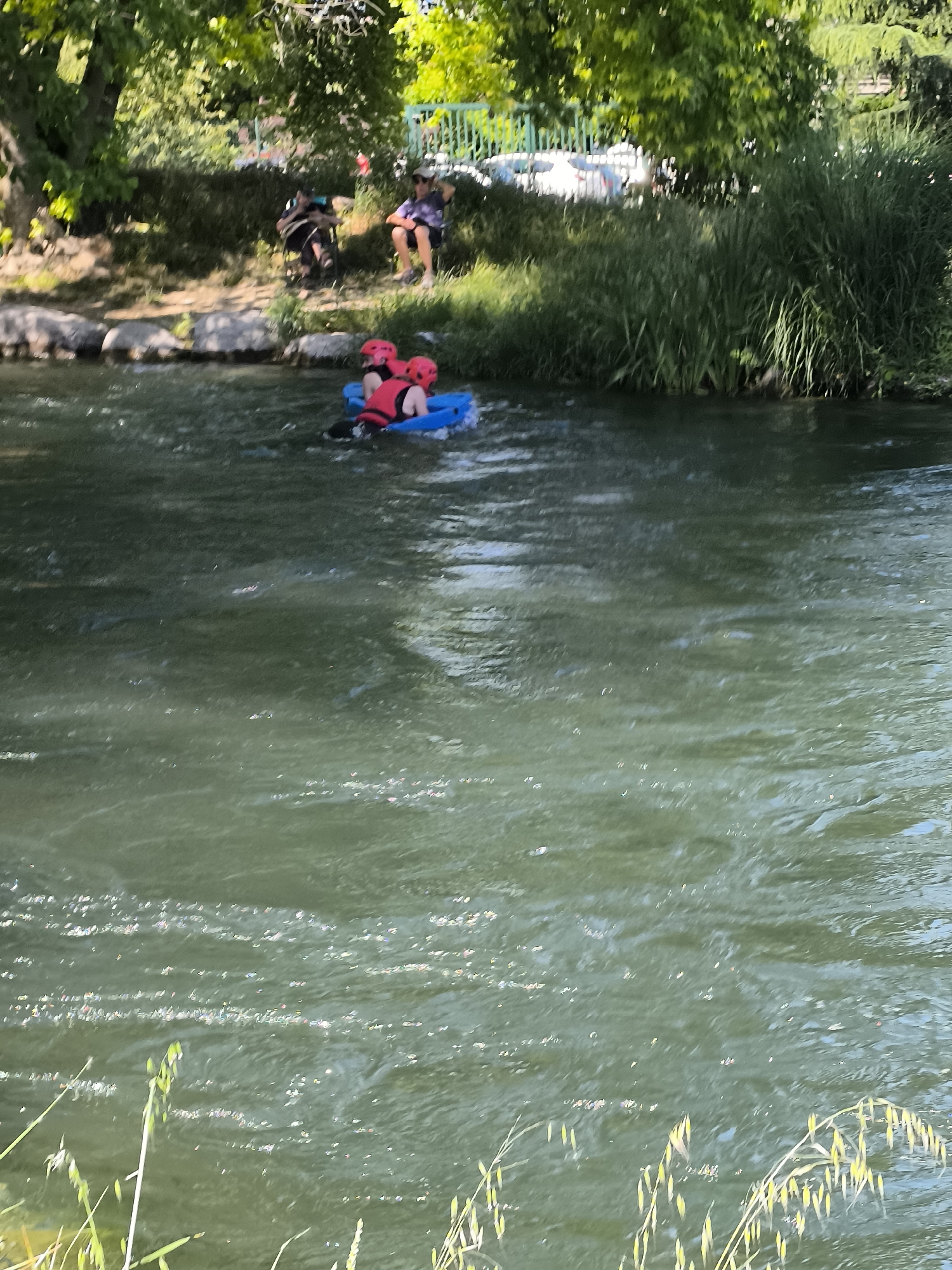 The image depicts two individuals in a blue inflatable raft, navigating a flowing river. Both individuals are wearing red helmets and life vests. The water is green, suggesting the presence of algae or sediment. On the riverbank, there are trees and grass. Two more individuals are visible on the riverbank. Based on the location, it can be stated that the scene is Saint-Pierre-de-Bœuf, France.