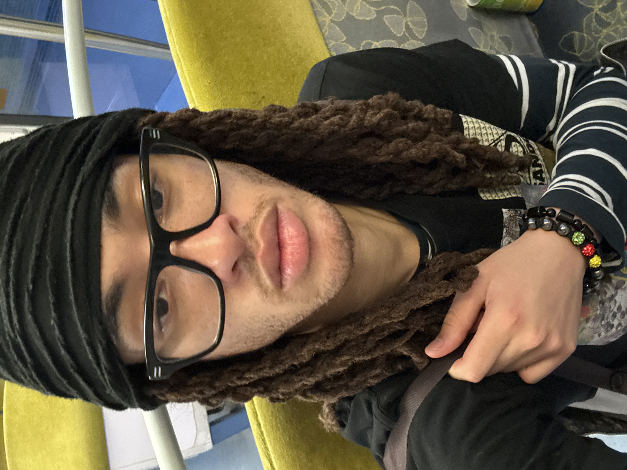 The image, rotated 90 degrees clockwise, presents a close-up portrait of a person inside what appears to be a form of public transport. The subject, a male, looks directly at the camera with a neutral expression. He wears black-framed glasses, distinguished by two small white horizontal lines on the temples. His hair is styled in dark brown dreadlocks, and he has light stubble on his chin and a mustache. A black, ribbed head covering or scarf is worn over his hair.
He is dressed in a black t-shirt featuring a faded, light-colored graphic design on the chest, layered over or with a long-sleeved shirt that has prominent black and white horizontal stripes. A dark jacket or piece of clothing is also visible. On his right wrist, he wears a bracelet made of black beads with additional colorful round beads in red, yellow, and green. His left hand rests on a dark strap or bag.
The background suggests an indoor setting with natural light. To the left and right, parts of a yellow or chartreuse-colored seat are visible. Behind him, another surface features a pattern of repeating grey abstract shapes on a darker grey background. Through a window, a clear blue sky and a white vertical pole are visible, indicating daytime. A partially obscured can, with visible green, white, and red sections, is in the upper right. The location is consistent with public transport in Orsay, France.