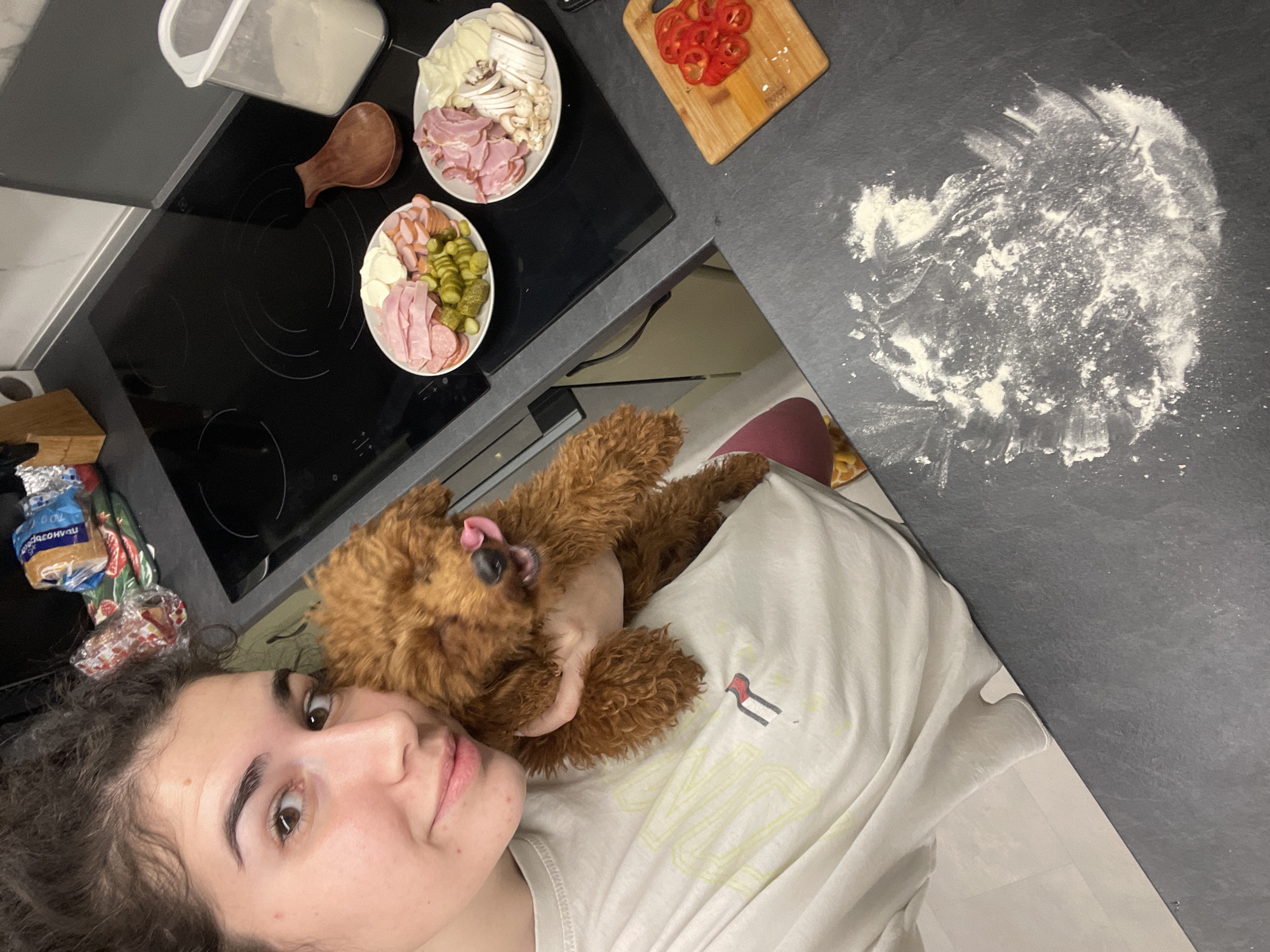 A high-angle shot shows a young woman taking a selfie with a fluffy brown poodle on her chest in a kitchen. The woman has curly dark hair and is wearing a white t-shirt. The poodle is licking its nose. A black stovetop is visible behind them, with a container, a wooden spoon, and two plates of pizza toppings on top. One plate contains sliced ham, mushrooms, and onions, while the other plate contains sliced ham, pickles, and sausages. A wooden cutting board with sliced red peppers is also visible. To the right of the stovetop, on a dark gray countertop, there is a patch of flour, suggesting someone has been preparing dough. The location is a kitchen, but specific details to determine if it's in Sofia, Bulgaria, are absent.