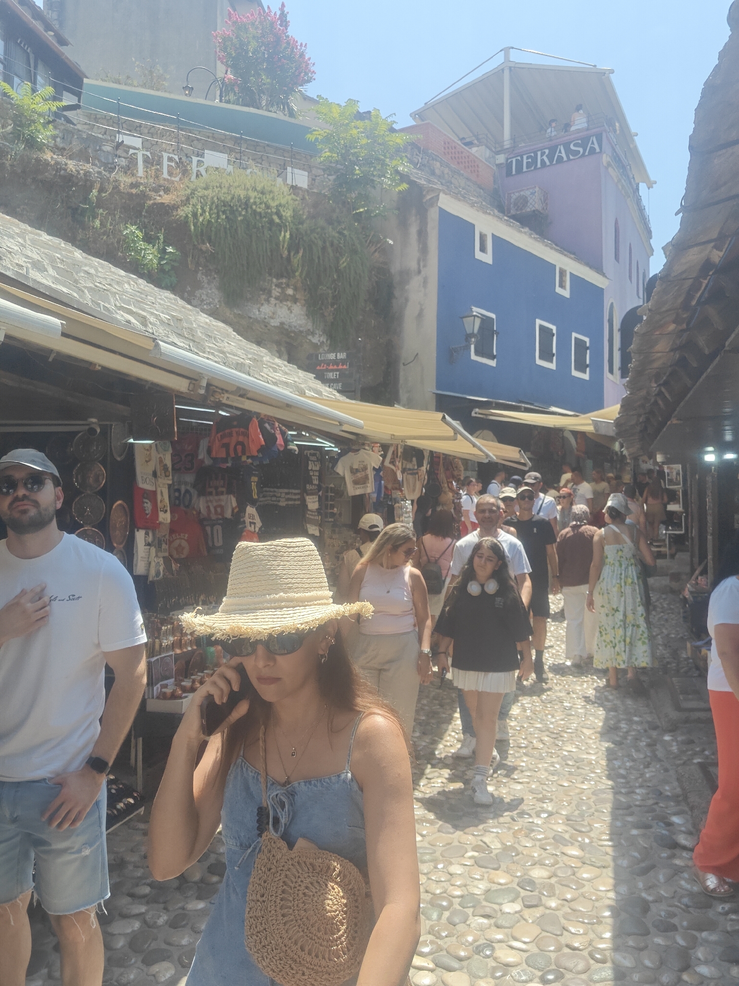 The image captures a bustling street scene in the old town of Mostar, Bosnia and Herzegovina, likely near the famous Stari Most (Old Bridge). A narrow cobblestone street is lined with shops displaying various souvenirs and apparel. In the foreground, a woman wearing a straw hat and a denim dress is talking on her mobile phone. To her left, a man in a white t-shirt and denim shorts stands with his hand on his chest. A crowd of tourists, dressed in summer attire, walks along the street, some browsing the shops. In the background, buildings with colorful facades, including a distinctive blue and purple one with the word "TERASA" visible, ascend a hillside. Sunlight casts shadows across the cobblestones, indicating a bright, sunny day. The overall atmosphere suggests a popular tourist destination.