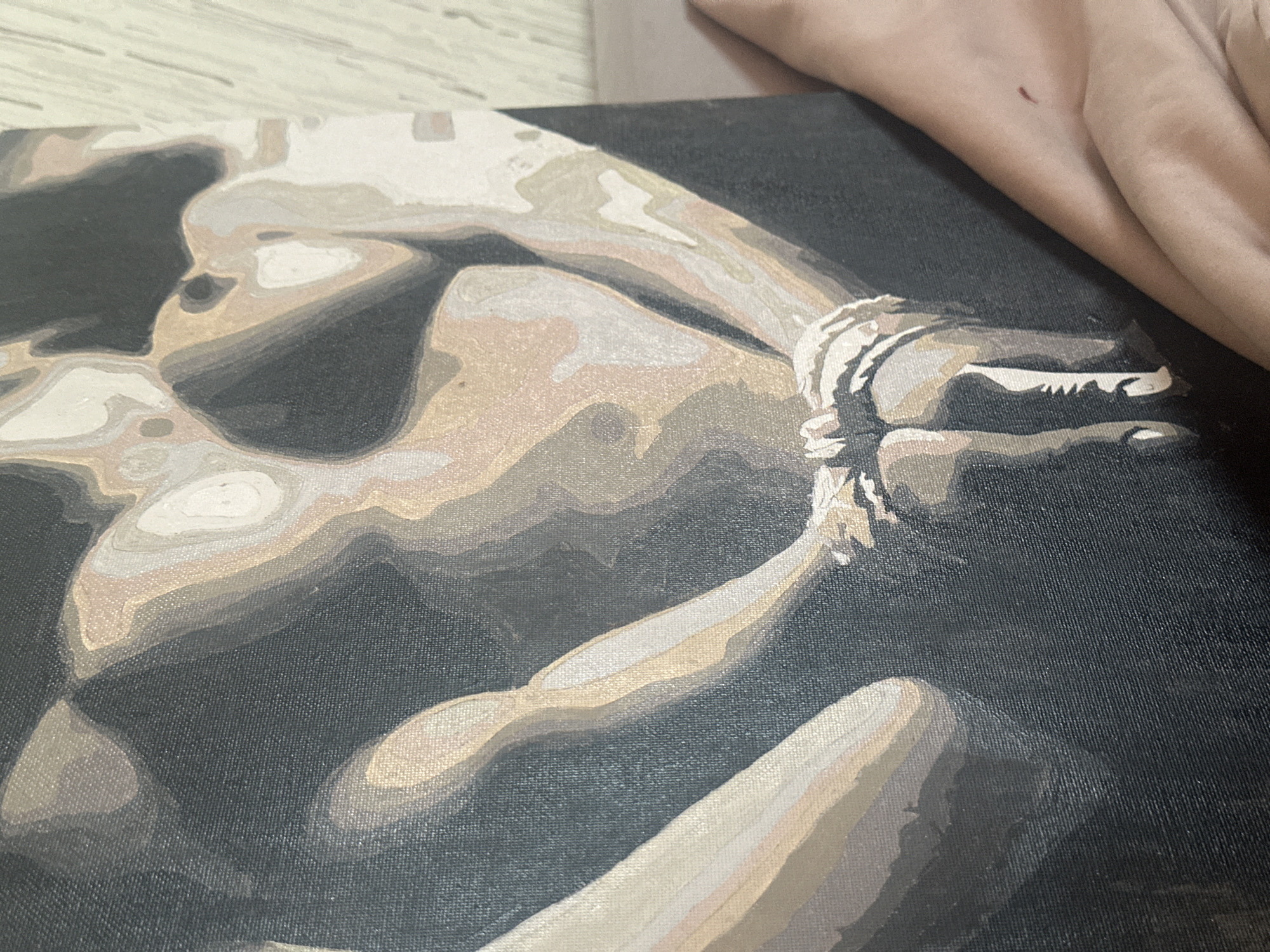 The image shows a close-up of a painting on canvas, depicting a nude figure in tones of brown, tan, and grey against a dark background. The figure appears to be reclining, with one arm bent and seemingly tied at the wrist. Details of the body are rendered in segmented blocks of color, typical of a "paint by numbers" style. Part of a human hand is visible in the upper right corner of the image, suggesting the person holding or displaying the painting. The canvas has a visible texture. The city of Высокий, Russia is not depicted in the image.