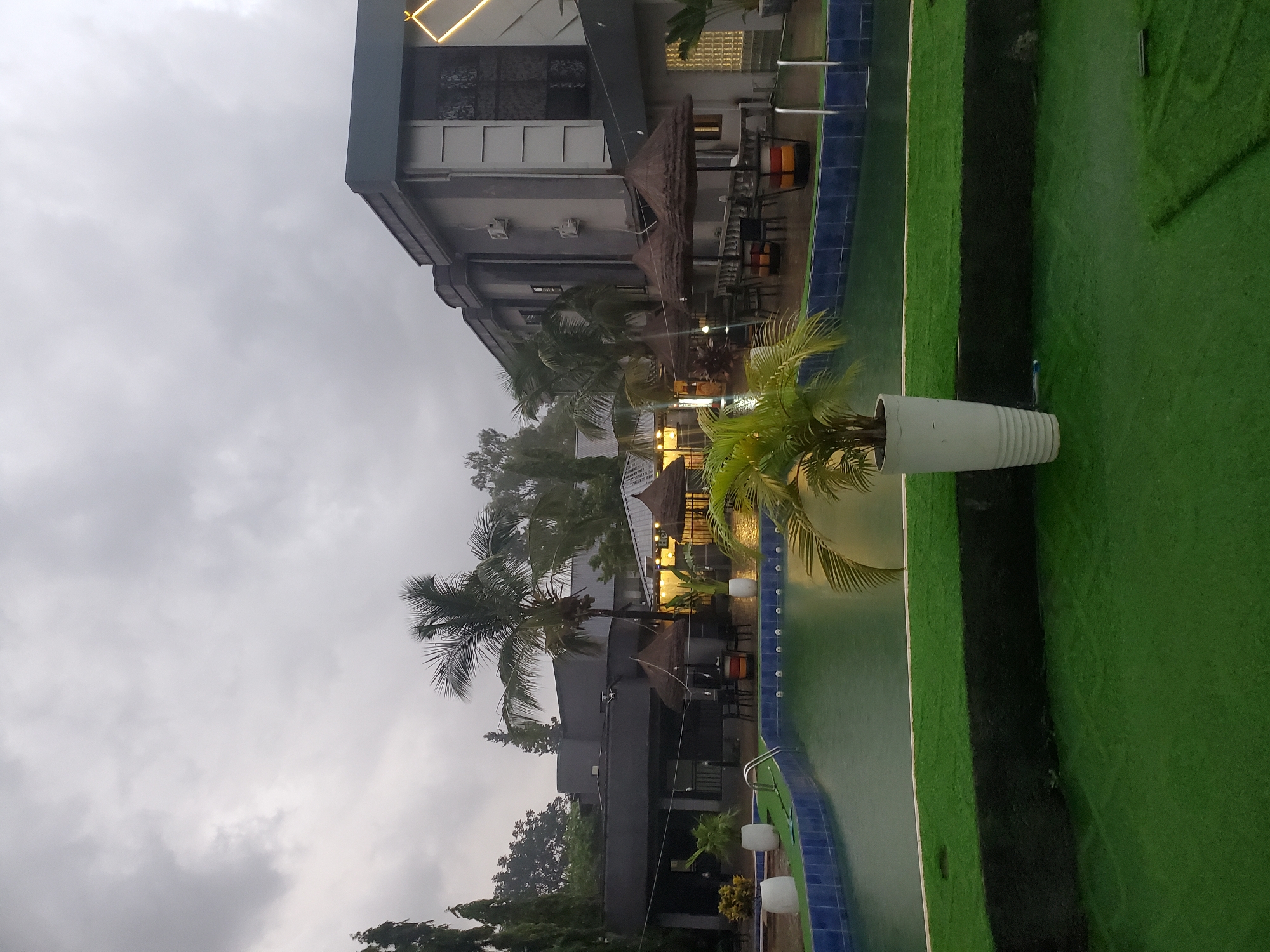 The image shows an outdoor scene with a building and a swimming pool. The building appears to be a hotel or resort, with multiple stories and a modern design. There are several thatched-roof structures that might be outdoor seating areas or cabanas. Palm trees and other tropical foliage are visible, suggesting a warm climate. The swimming pool is a prominent feature, with blue tiled edges and what appears to be artificial turf surrounding it.
There are no people visible in the image. The objects present include the building, furniture (chairs and tables), umbrellas or shades, planters with plants, and the swimming pool itself.
The sky is overcast with gray clouds, indicating it might be a cloudy or rainy day. The overall lighting suggests it could be daytime, but the lack of direct sunlight and the diffused light make it difficult to determine the exact time. The presence of lush greenery and palm trees points to a tropical or subtropical location. Given the location context provided, this could be a resort in Jos, Nigeria.
There is no visible text in the image.
The scene appears to be serene and possibly a bit gloomy due to the weather. The setting is that of a leisure or hospitality establishment designed for relaxation and outdoor enjoyment.