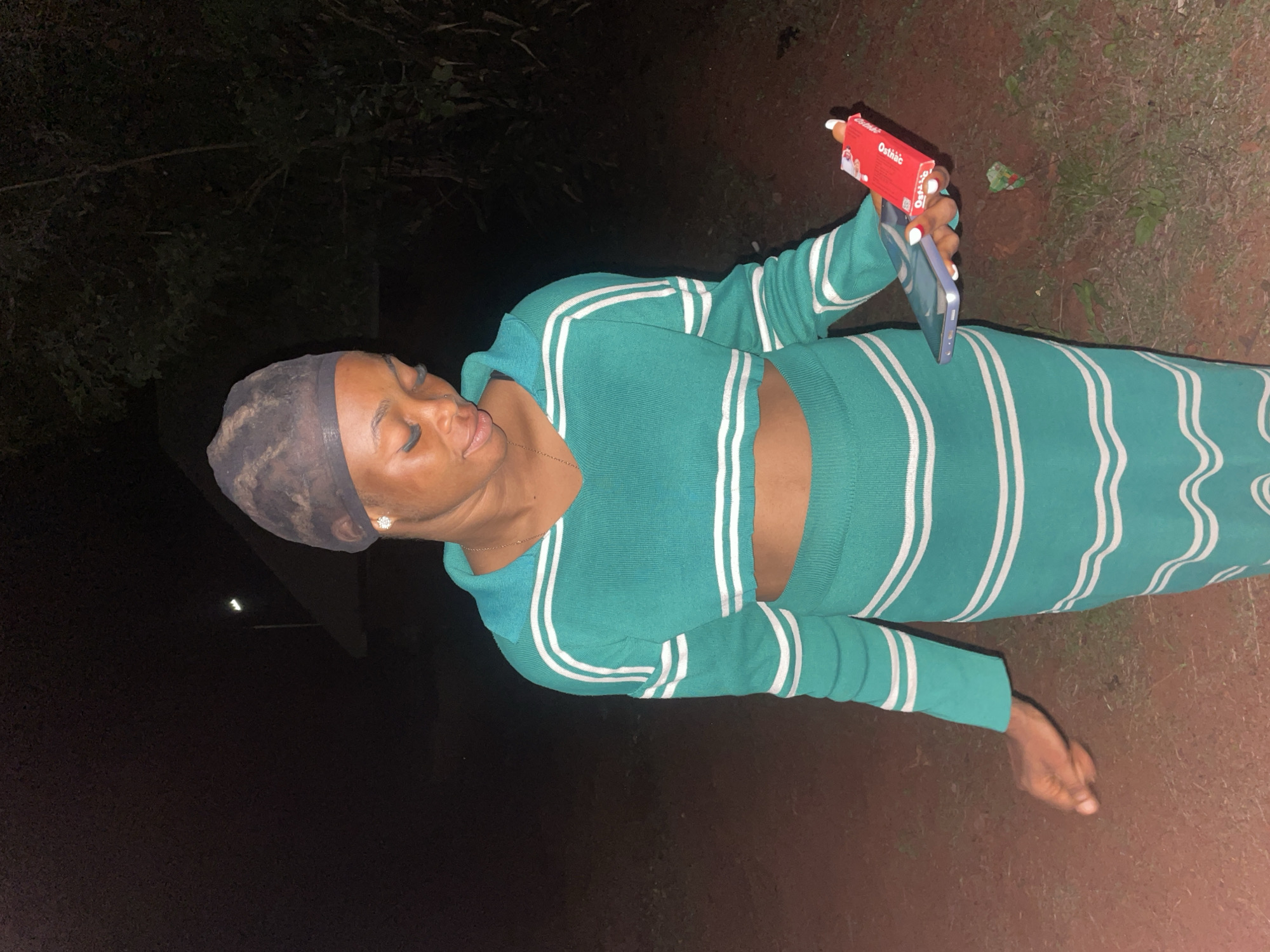 The image depicts a woman in Ogbe, Nigeria. She is wearing a light-green, two-piece outfit with white stripes and a dark-colored head covering. She holds a blue cell phone in one hand and a small red box labeled "Ostinac" in the other. The background is dark, suggesting it is nighttime. The ground is a reddish-brown color, and some green vegetation is visible in the upper right. The lighting is poor, with a single light source illuminating the woman.