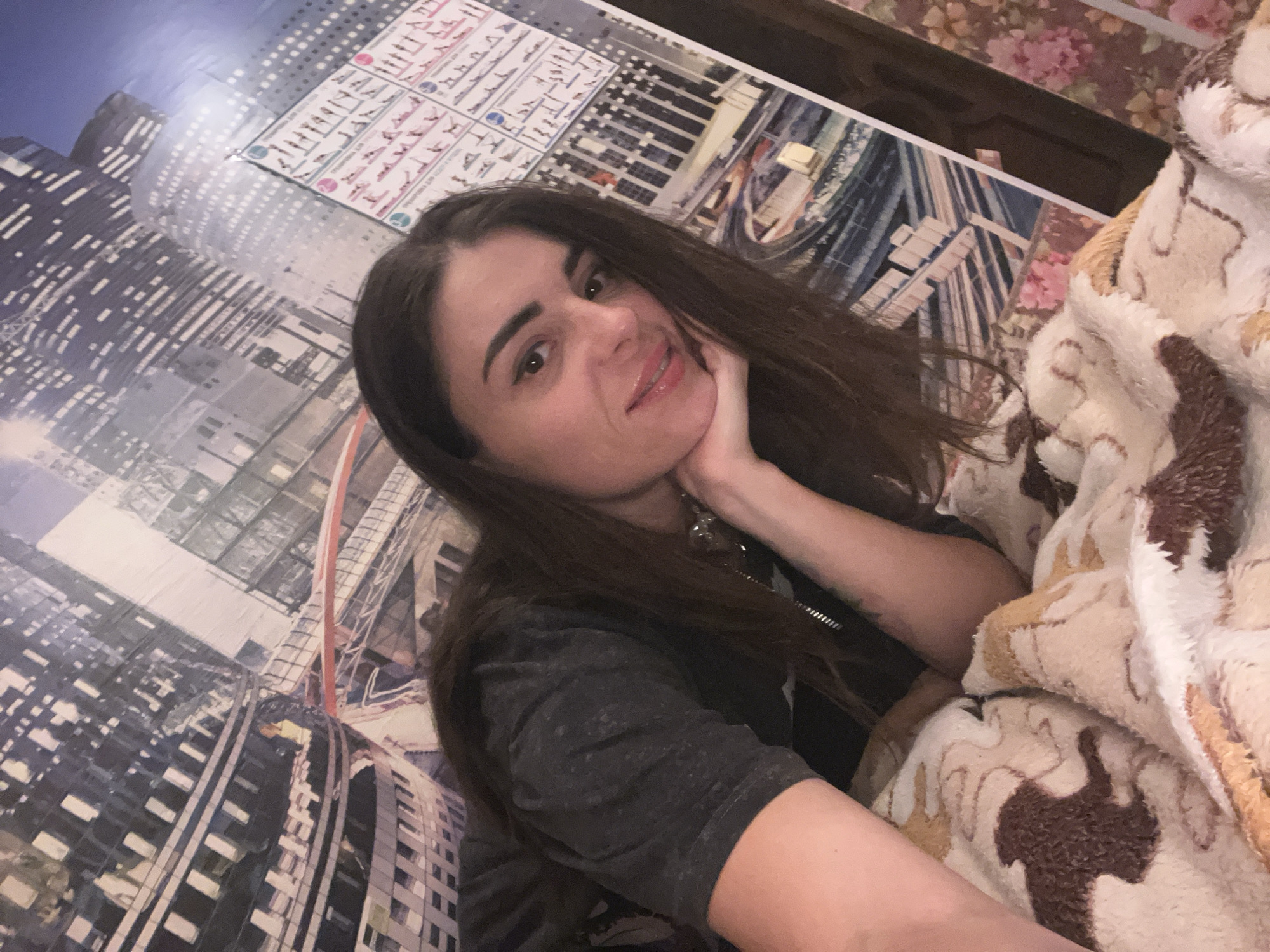 The media file is a selfie of a woman with long, dark hair and brown eyes. She is smiling and resting her chin on her hand. She is wearing a dark t-shirt. The background appears to be a wall with a large mural depicting a city skyline with skyscrapers. There is also a poster on the wall with figures demonstrating various yoga poses. A patterned blanket is visible in the foreground, partially covering the woman. The lighting suggests it is indoors, possibly daytime. The woman's expression appears relaxed and content. There is no visible text other than what appears to be instructions or labels on the yoga poster.