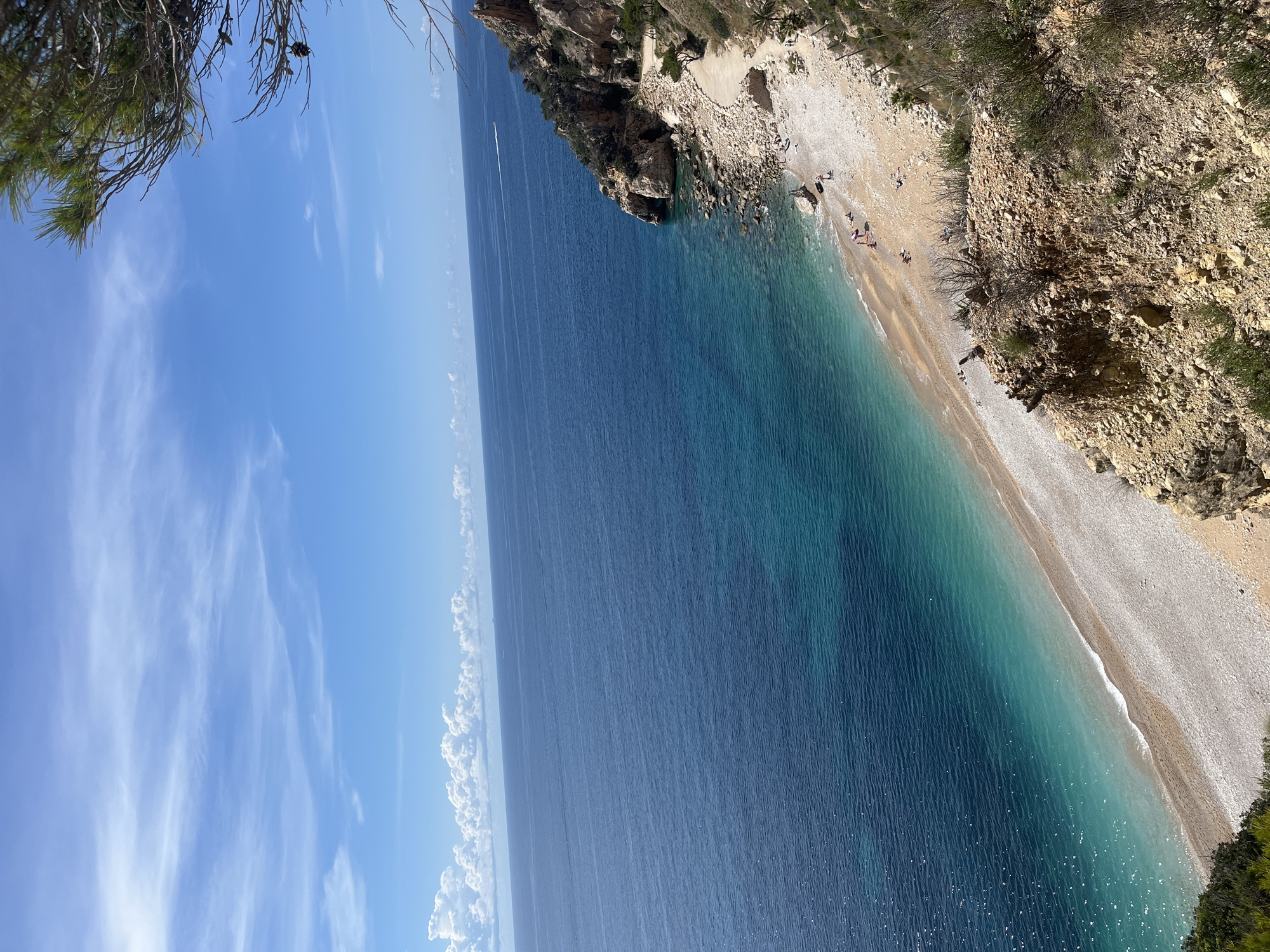 This image captures a view of a beach at el Poble Nou de Benitatxell / Benitachell, Spain. The perspective is from a high vantage point, looking down at the coastline. The beach is composed of pebbles and sand. The water varies in color from deep blue to turquoise. Several people are seen on the beach. Rocky cliffs edge the shoreline. The sky is blue with scattered clouds. Vegetation is visible on the cliffs.