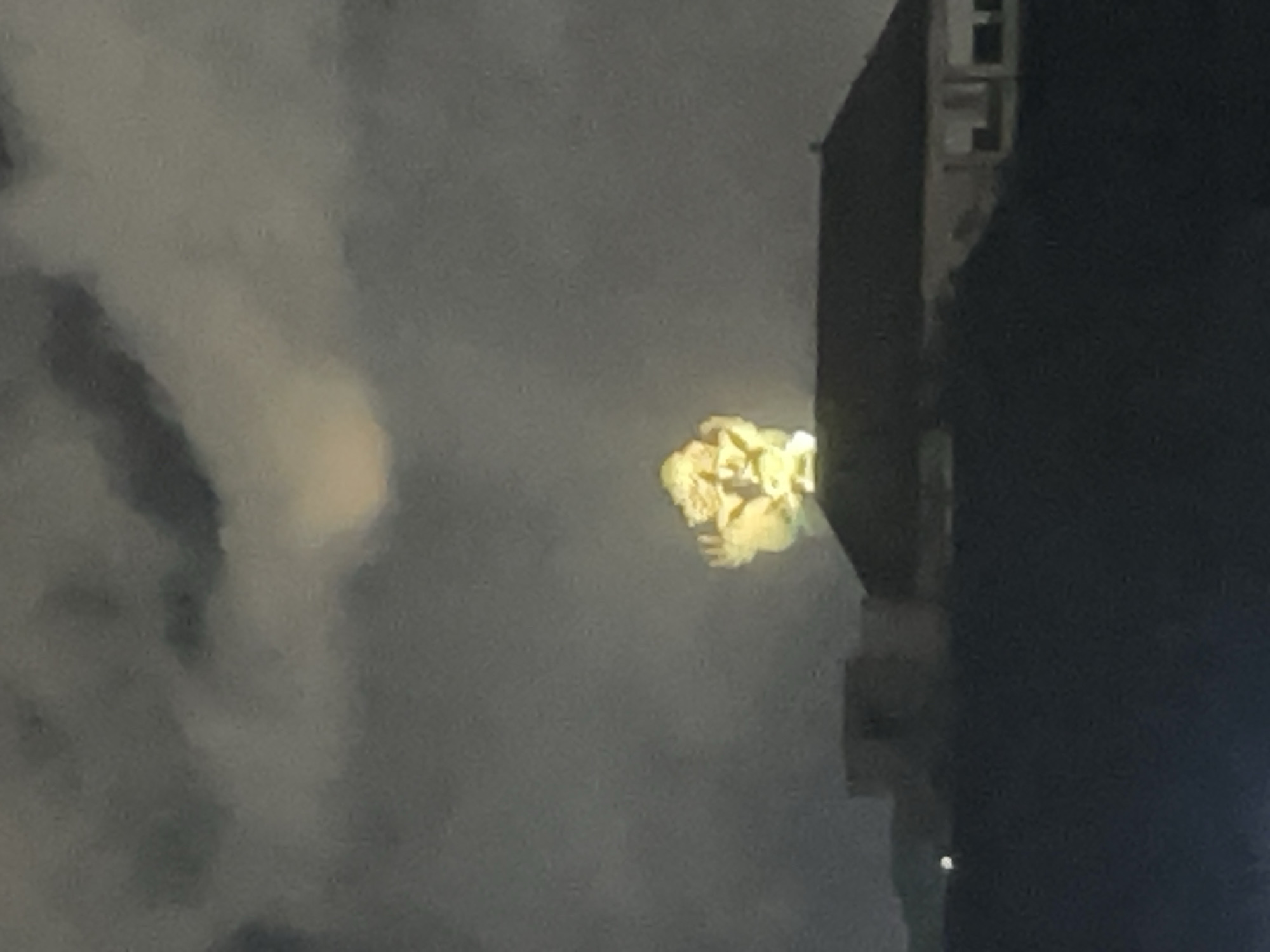 A brightly lit statue, possibly a deity, is visible above a building in Kuta Selatan, Indonesia, at night. The statue is illuminated from below and appears to be made of stone or a similar material. It is silhouetted against a cloudy sky with a faint glow in the background, potentially from the moon. The building below the statue is dark, with its roofline visible. Further in the foreground, another building is dimly lit. The overall scene is dark and atmospheric, with the illuminated statue serving as the focal point.