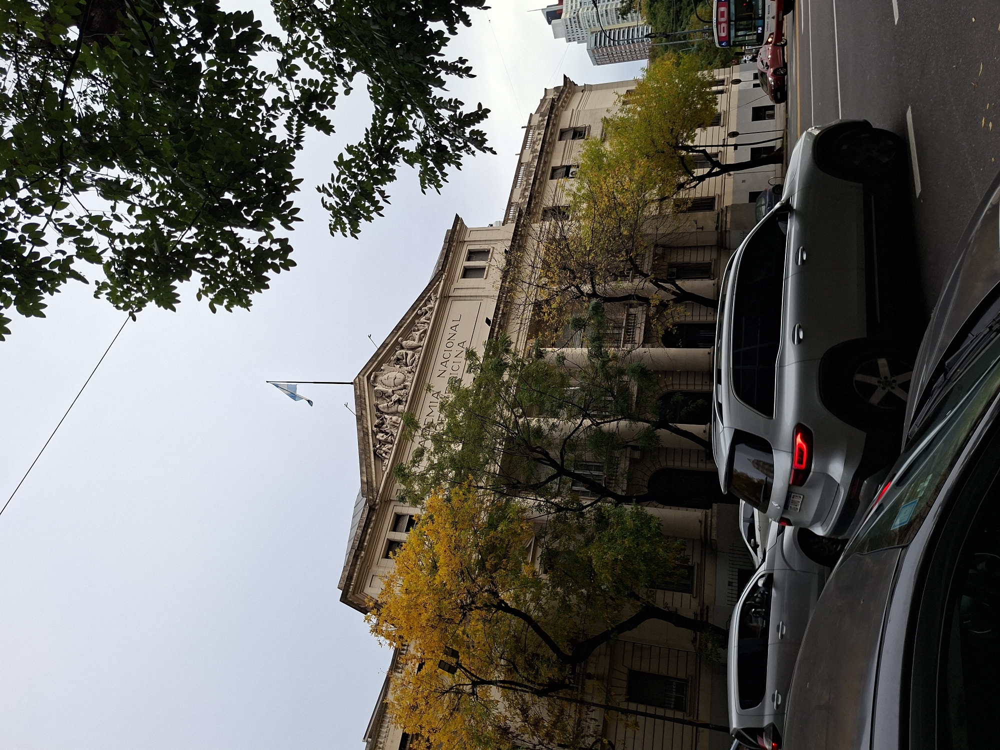 The image captures a street scene in Buenos Aires, Argentina, featuring the Academia Nacional de Medicina building. The building, constructed in a neoclassical style, has an Argentine flag atop its roof. In the foreground, multiple cars are parked alongside the street. Trees frame the building, with some displaying yellow leaves, suggesting it's autumn. Additional buildings are visible in the background.