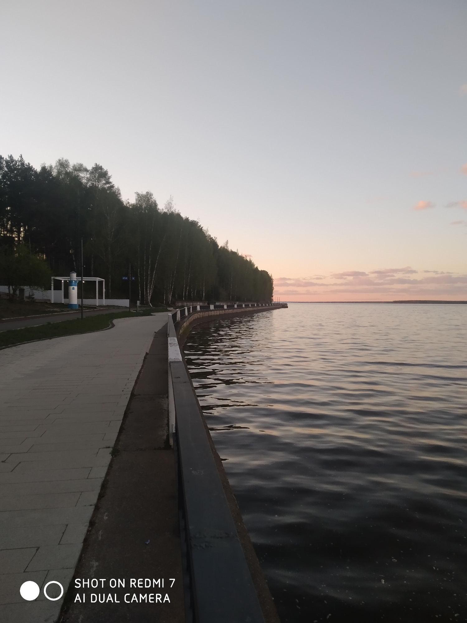 The image depicts a waterfront view at dusk in Chaikovsky, Russia. A paved walkway with a low concrete barrier runs along the edge of the water, with trees bordering the path on the left. The water stretches into the distance under a pale sky, reflecting the muted colors of the sunset. A white and blue decorative lighthouse-like structure is visible along the walkway. The scene appears calm and still.