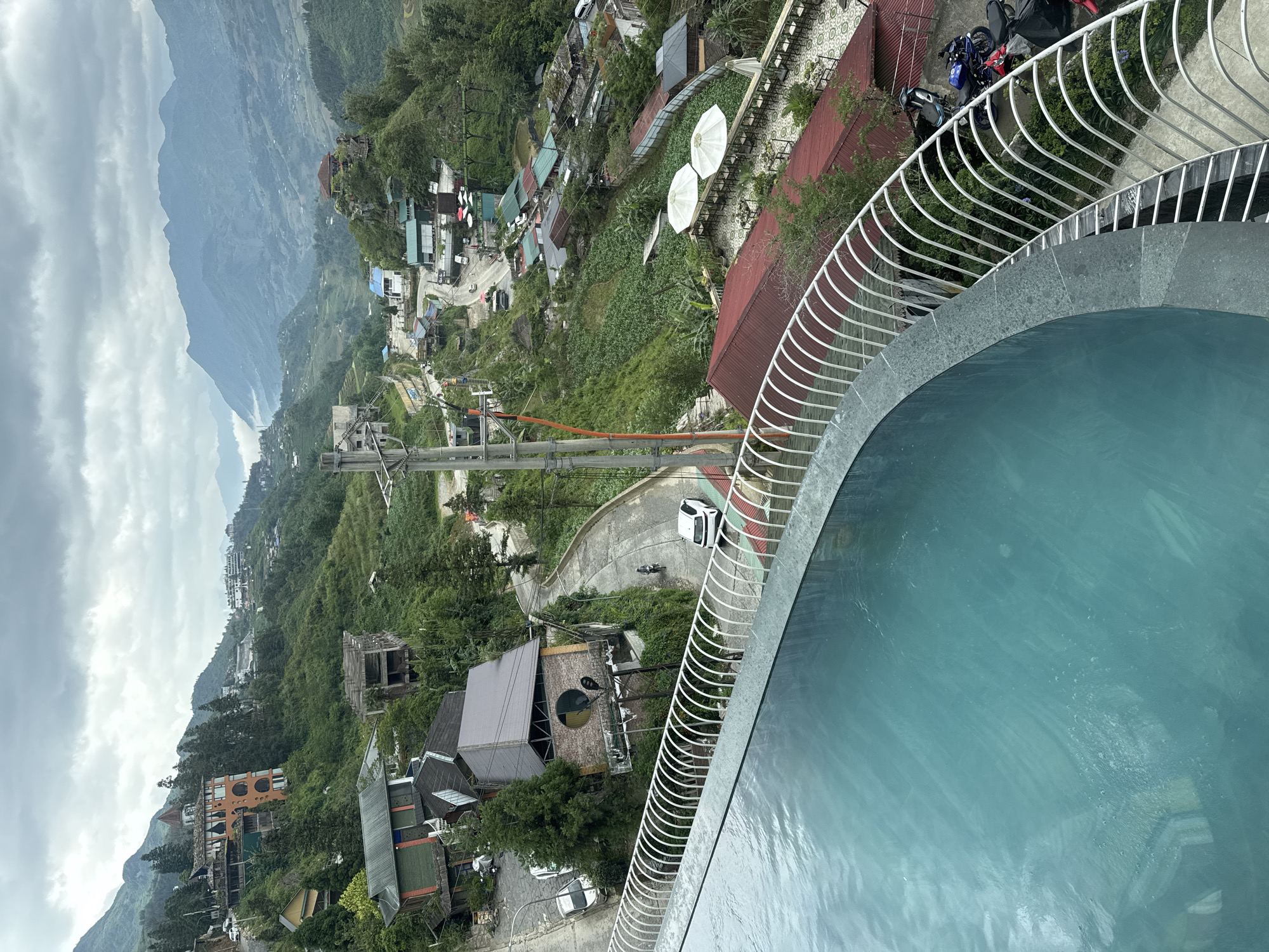 A high-angle, slightly tilted view looks down on a landscape featuring a swimming pool, buildings, and distant mountains, likely in Sa Pa Ward, Vietnam. The foreground is dominated by a curved swimming pool with light blue water, bordered by a white metal railing and a stone or concrete rim. Beyond the pool, the scene opens to a landscape of buildings nestled among green vegetation. Some buildings have red roofs, while others have gray or brown. A road winds through the settlement, with a white car and a motorcycle visible on the road. In the background, hazy mountains are visible under a cloudy sky. Power lines and poles are also visible in the midground, stretching across the landscape. Several satellite dishes can be seen on a building.