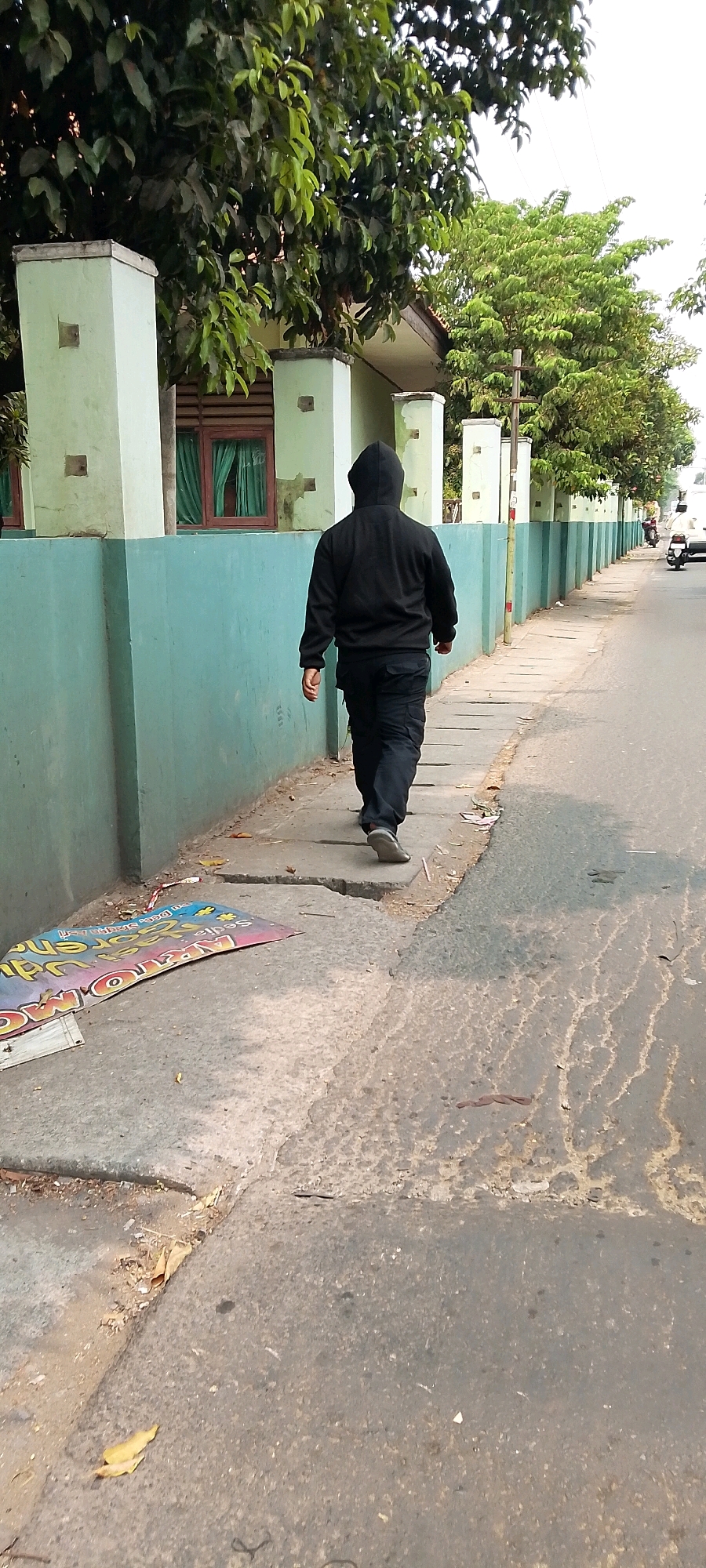 A person wearing a black hooded jacket and black pants walks on a sidewalk next to a turquoise wall in Bekasi, Indonesia. The sidewalk is made of concrete slabs and has some debris on it, including fallen leaves and a discarded colorful sign that reads "AKAR MO" and other illegible text. Beyond the wall, a building with green trim and windows can be seen, surrounded by trees. The street to the right of the sidewalk is paved and appears to be a road with a motorcycle in the distance. The sky is overcast.