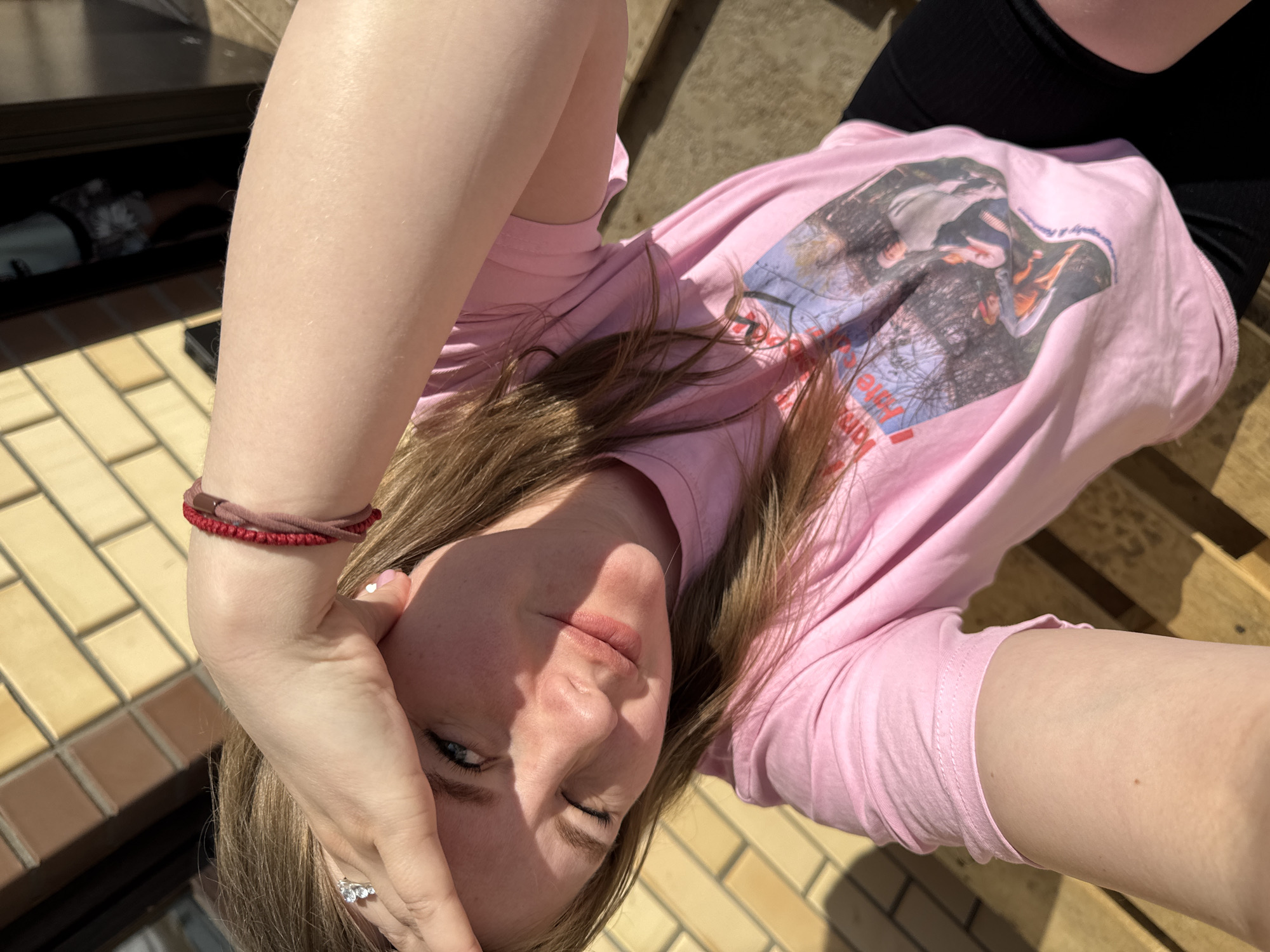 An upward-angled, close-up photograph captures a young fair-skinned woman in an outdoor setting in the city of Gornyi (Dvurechenskoe S/P), Russia. The woman, with long light brown hair, is positioned with strong sunlight illuminating her from the left, casting shadows across her face. Her left hand is raised across her forehead, shielding her eyes from the sun. A silver-colored ring with multiple stones is visible on the ring finger of her left hand. Her left eye, appearing blue, is open, while her right eye is partially squinted. She is wearing a pink short-sleeved t-shirt featuring a graphic print of several individuals in a natural, tree-lined setting, and black ribbed shorts. On her right wrist, a red braided bracelet and a maroon fabric bracelet are visible. The background consists of a light yellow brick wall laid in a stretcher bond pattern, with a dark doorway or window reflection visible behind her right shoulder. Below her, wooden planks or steps are discernible.