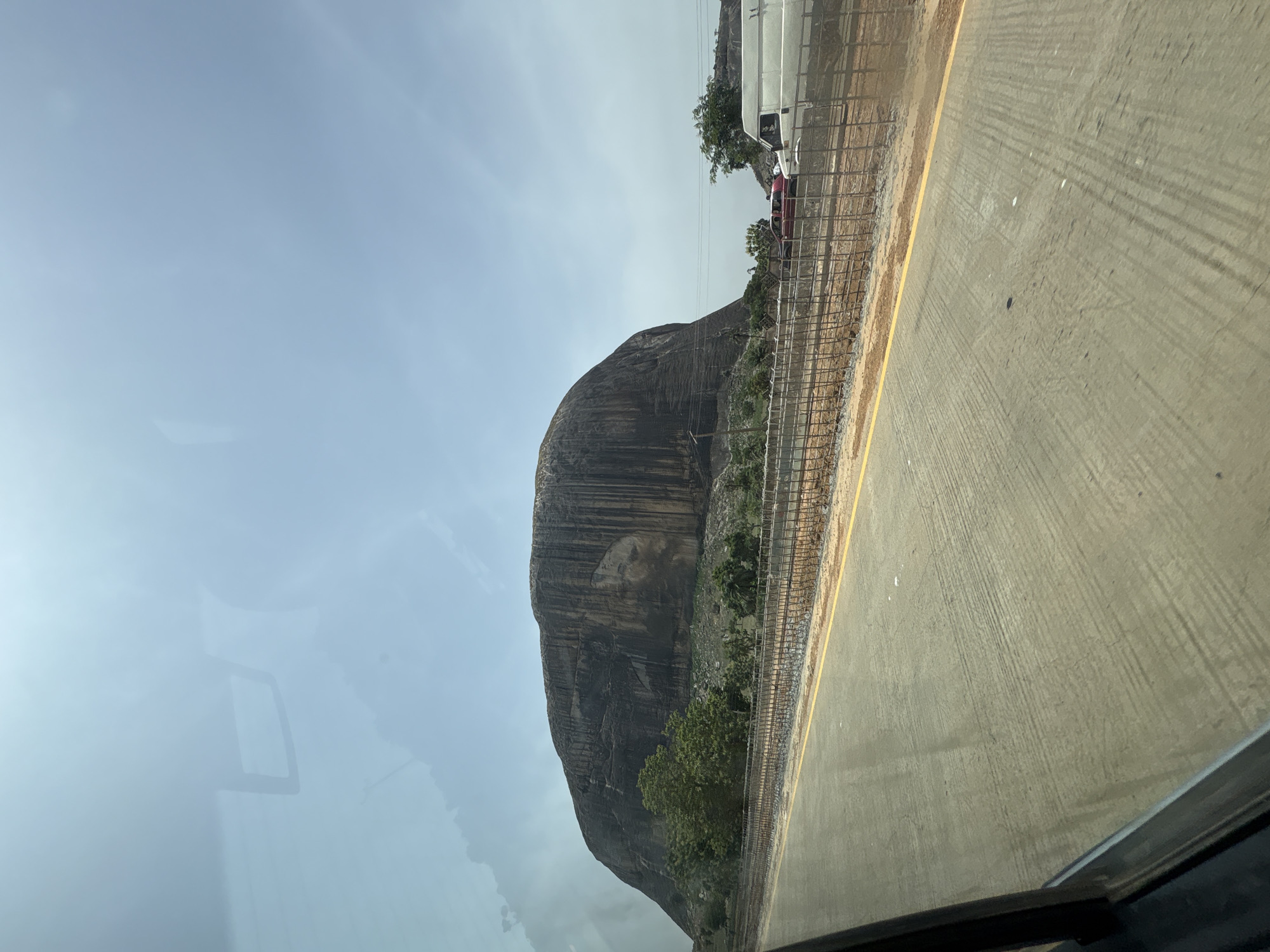 The image shows Zuma Rock, a large monolith, as seen from a vehicle traveling on a road in Gwagwalada, Nigeria. The rock face is prominently featured to the left. A road with a yellow painted line runs along the right side of the frame, with a metal fence separating the road from the base of Zuma Rock. Other vehicles can be seen in the background. The sky is overcast. The view is slightly distorted due to being seen through a vehicle window.
