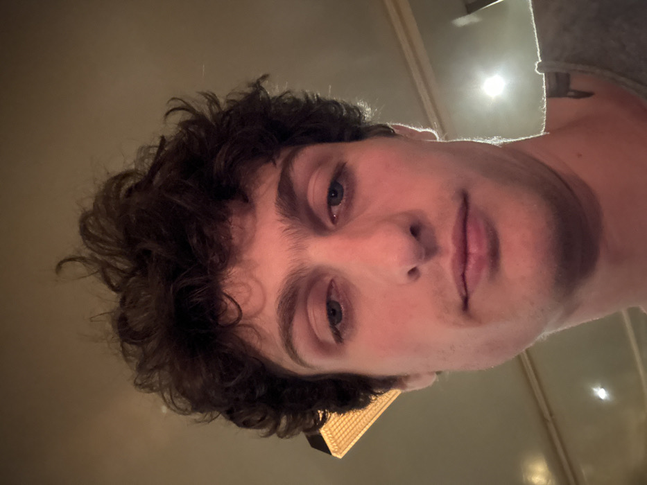 A slightly downward-angled selfie of a fair-skinned young man with curly, dark hair. His blue eyes look directly at the camera. The background consists of a dimly lit interior with a visible ceiling fixture and a glimpse of a light source reflecting off a wall. A gray shoulder appears in the upper right corner. The man's facial expression is neutral, and the overall lighting is soft.
