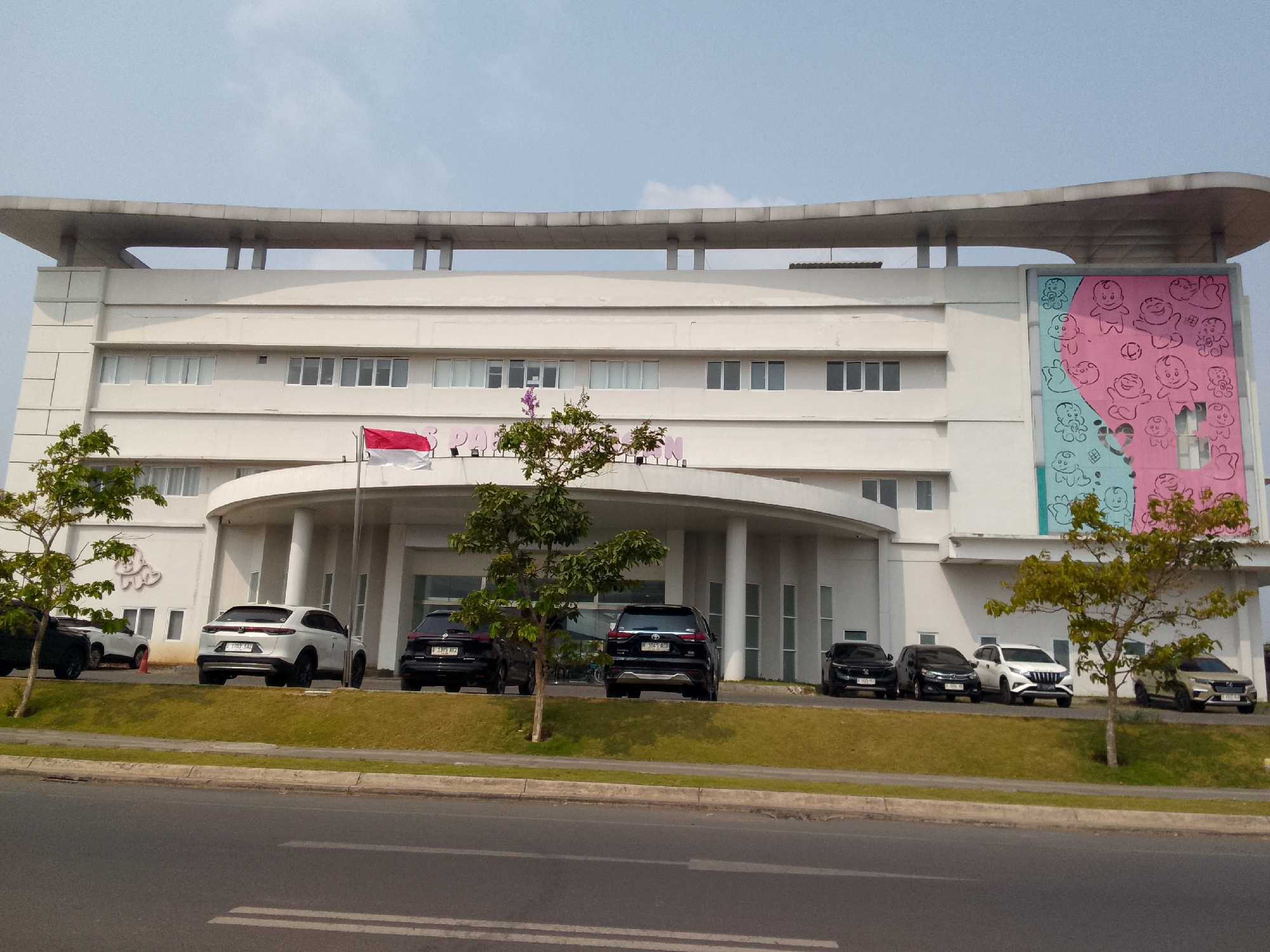 The image depicts the facade of a multi-story building with a distinctive curved roofline, situated in Jayamekar, Indonesia. The building is primarily white, with numerous evenly spaced rectangular windows on its upper floors. A large, colorful mural featuring cartoonish drawings of babies adorns the right side of the building's exterior. In front of the building, a row of parked cars, including a white SUV and several dark-colored SUVs and sedans, are visible on a manicured grassy area. A flag, appearing to be the Indonesian flag (red and white), is displayed on a flagpole near the entrance. Several trees with green foliage are planted in front of the building, and a road with white lane markings runs across the foreground. The sky above is clear with a few wispy clouds.