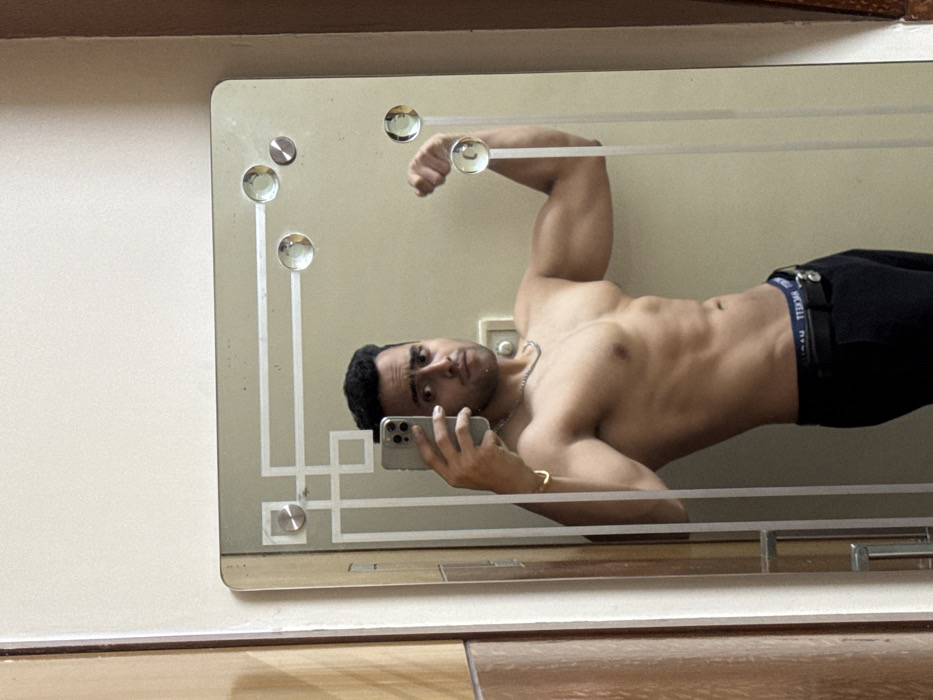 A shirtless man poses in a mirror, flexing his bicep. He holds a smartphone in his left hand, presumably taking the photo. He wears dark pants and a silver necklace. The mirror has a decorative frame with circular embellishments and is mounted on a wall with visible wood trim at the top and a tiled surface below.