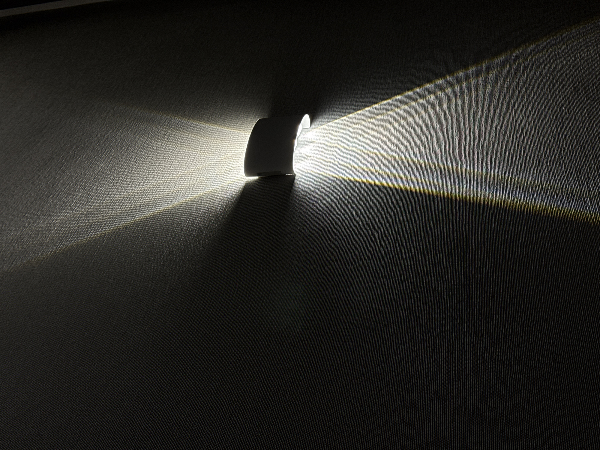 A cylindrical, white wall light is mounted on a dark, textured wall. The light emits two beams of light, one diverging to the left and the other to the right, creating contrasting areas of light and shadow on the wall's surface. The environment appears dimly lit, emphasizing the light emanating from the fixture. The location and city are not visible in the image.