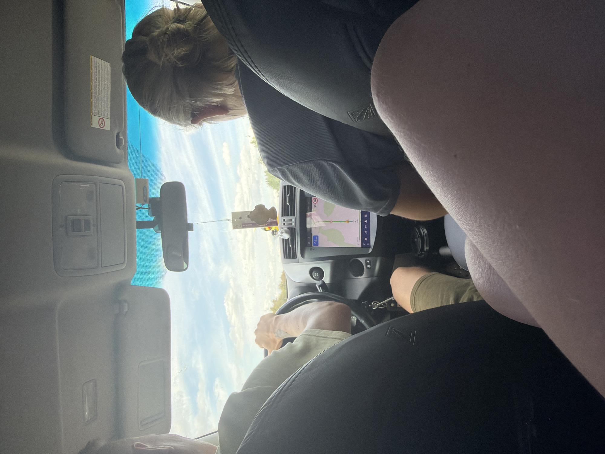 The image shows the interior of a car from a passenger's perspective. In the foreground, the legs of a person are visible. In the middle ground, a person is driving the car, their hands are on the steering wheel, and they are wearing a light-colored shirt and green shorts. The car's dashboard is visible, featuring a central screen displaying a navigation map. To the right of the driver, another person with blonde hair tied back is seated. The exterior view through the windshield shows a cloudy sky and trees. The overall scene depicts a journey inside a vehicle. The image does not contain any information that would suggest the location is in Kruglovka, Russia.